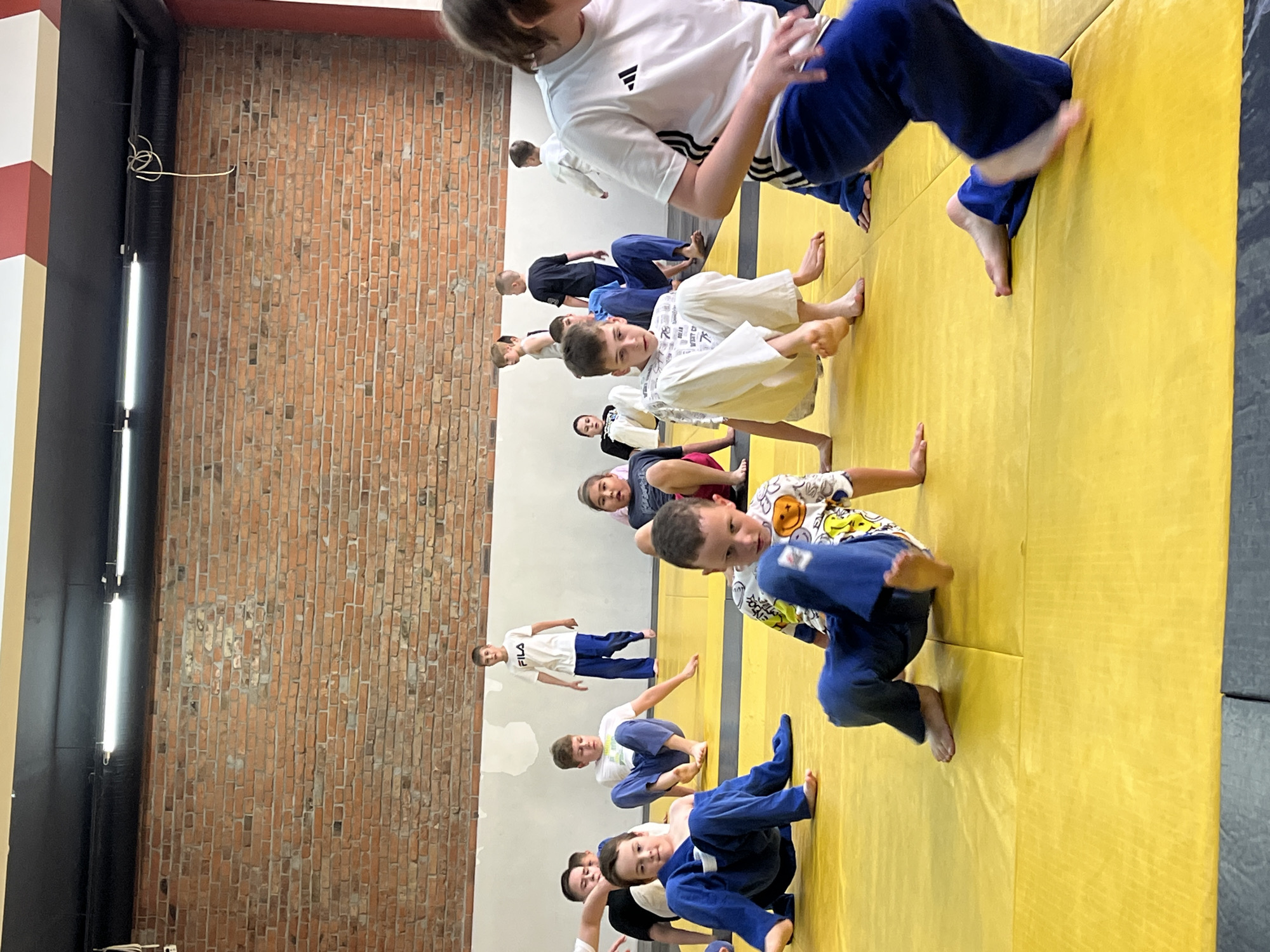 The image depicts a group of children in a dojo, likely participating in a judo or martial arts class. They are on a yellow mat, with a brick wall in the background. Several children are wearing traditional judo uniforms (gi), while others are in casual athletic wear. Some children are sitting or kneeling, while others are in motion, suggesting a training exercise or drill. The lighting appears to be from overhead fluorescent fixtures. The environment is consistent with an indoor sports facility. The image appears to have been taken in Krasnoyarsk, Russia, based on the provided location information.