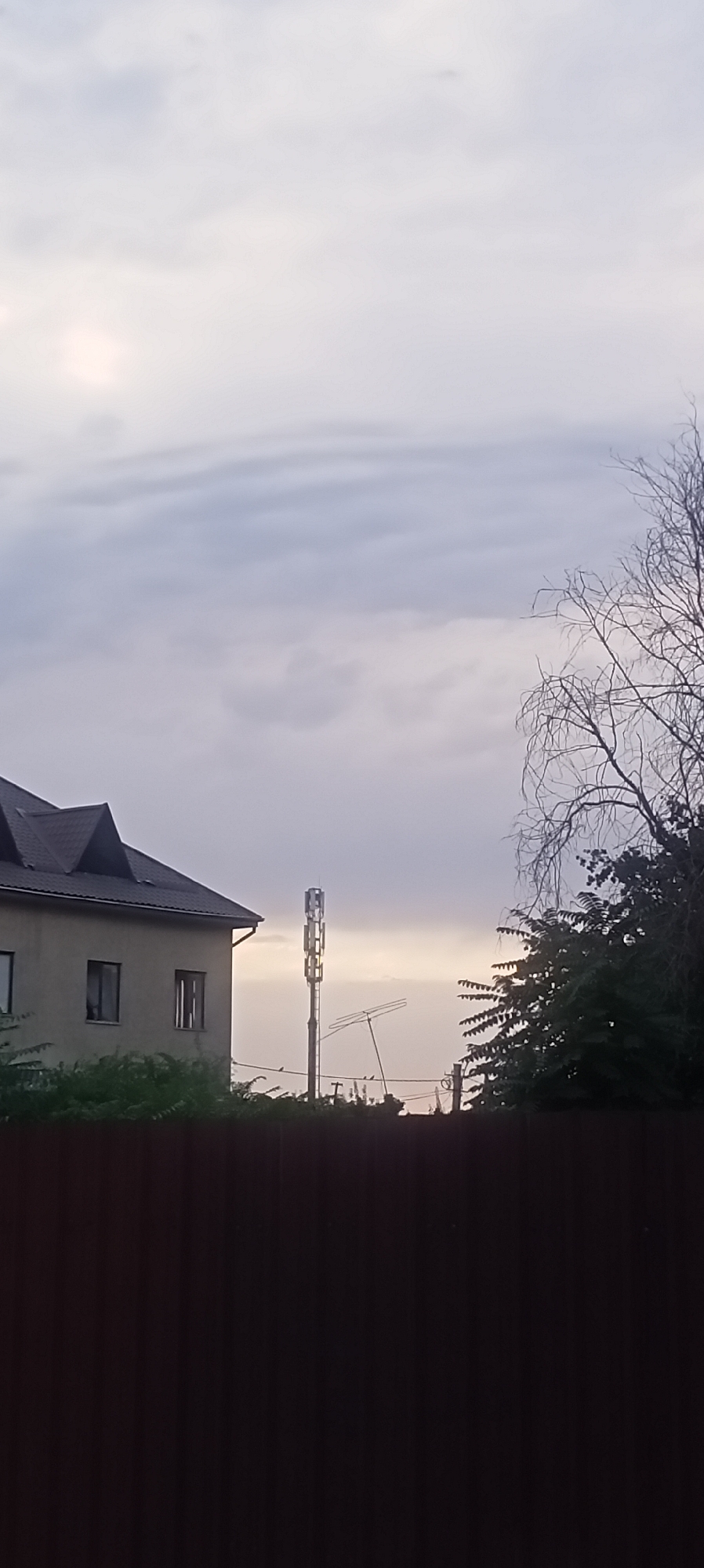 A telecommunications tower, some trees, and a building are visible against a cloudy sky. The tower is tall and metallic, with antennas at the top. The trees are green and leafy, with some bare branches visible on the right side of the image. The building is a light-colored, two-story structure with a dark roof. A dark brown fence runs along the bottom of the image. The overall scene suggests an urban or suburban setting.