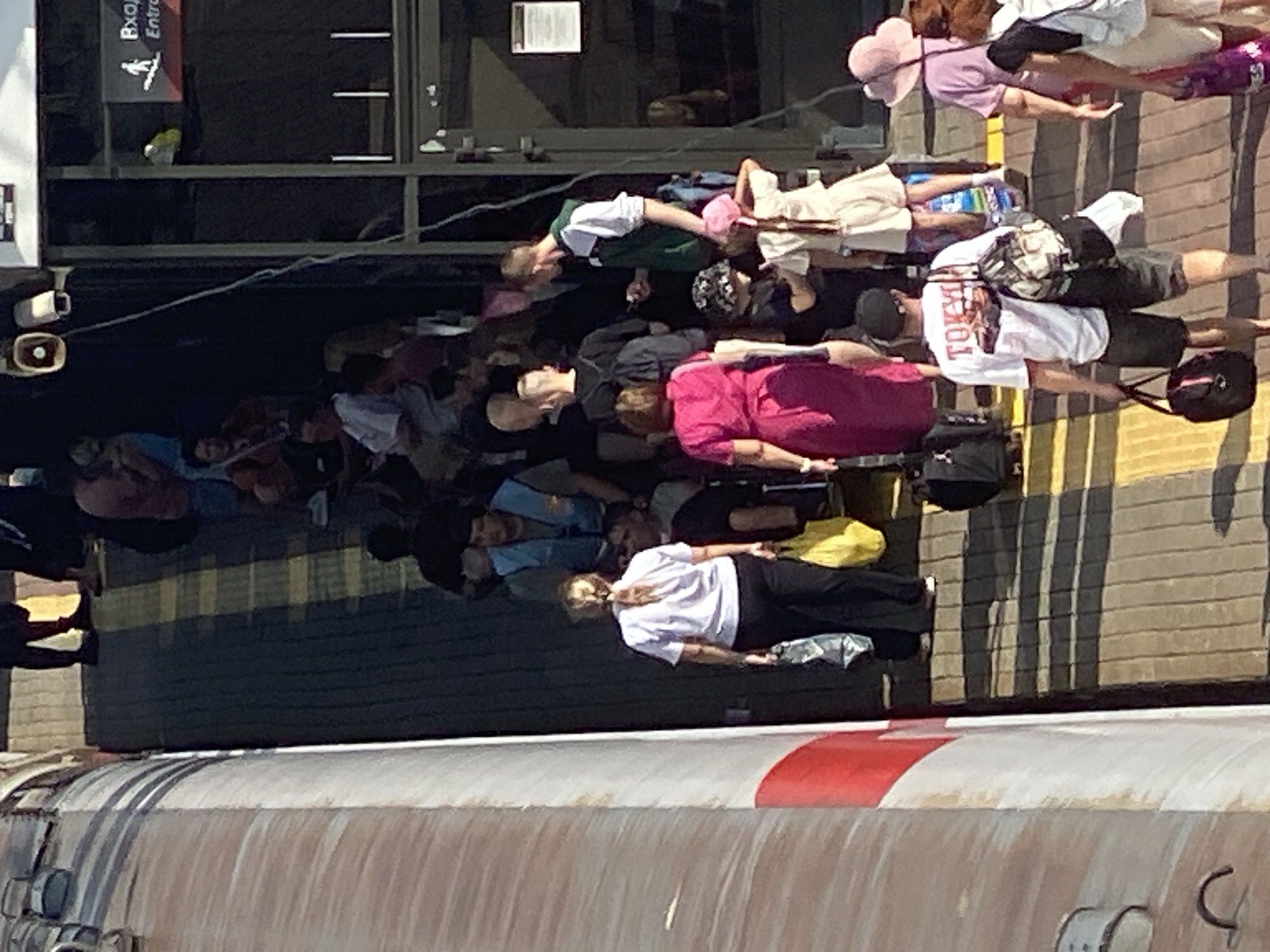 The image depicts a group of people on a ramp or walkway, possibly a ferry terminal or a similar transportation hub, with a large, textured structure on the left side that appears to be part of a vessel. The walkway is paved with a grid of yellow and dark gray tiles, with shadows casting diagonal stripes across the surface.
In the foreground, a woman with blonde hair in a ponytail, wearing a white polo shirt and black pants, is walking away from the camera, carrying a plastic bag in her left hand and a yellow shopping bag in her right. Behind her, a man wearing a white t-shirt with "TOKY" visible on the back, dark shorts, and a baseball cap is walking, carrying a duffel bag over his shoulder and a black gym bag in his left hand.
Further up the ramp, a woman in a bright pink dress is walking towards the camera, carrying a yellow shopping bag. Several other individuals are visible on the ramp, some standing and others walking. One man wears a blue vest over a grey shirt. Another individual, partially visible, appears to be a child wearing a hat and holding a phone. A person in a green and white jacket is standing and looking at a phone.
In the background, there are more people, and a building or structure with signage is visible. The overall scene suggests a public gathering or boarding area for some form of transport.
While the image is provided in the context of Novokuznetsk, Russia, there are no specific visual cues within the image itself that definitively place it in that city. The signage in the background is not clearly legible to confirm a specific location.