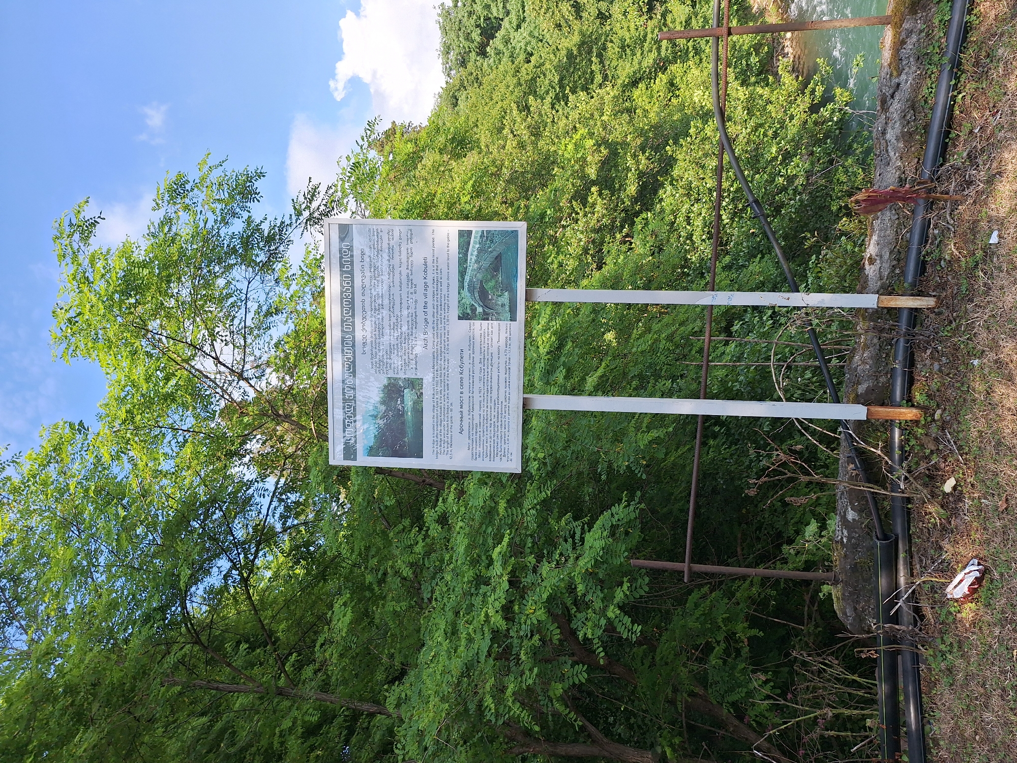 This is a medium shot, outdoor, eye-level photograph of a signpost in Kobuleti, Georgia, near a river. The signpost is white and has text and images in Georgian and English. The text describes the Arch Bridge of the village of Kobuleti. The sign is positioned in front of a dense green forest with a blue sky and white clouds visible above. A river is visible to the right of the signpost, with trees on its opposite bank. The foreground shows dry grass, some black pipes, and a discarded piece of red and white packaging. A rusty metal fence runs horizontally across the middle ground behind the signpost.