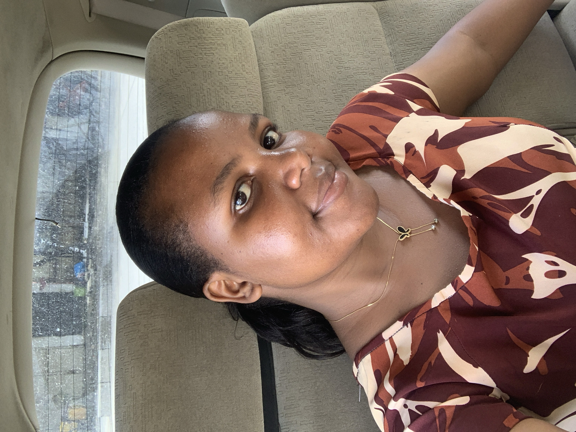 A woman is lying on the back seat of a car. She is facing upwards and looking directly at the camera. She has dark skin, short black hair, and is wearing a brown, white, and tan patterned shirt. A delicate gold necklace with a small butterfly pendant is visible around her neck. The car seat is upholstered in a light brown, textured fabric. Through the car window, a blurry urban landscape is visible. Based on the request, it can be inferred that this is a scene taken in Dar es-Salaam, Tanzania, inside a vehicle.