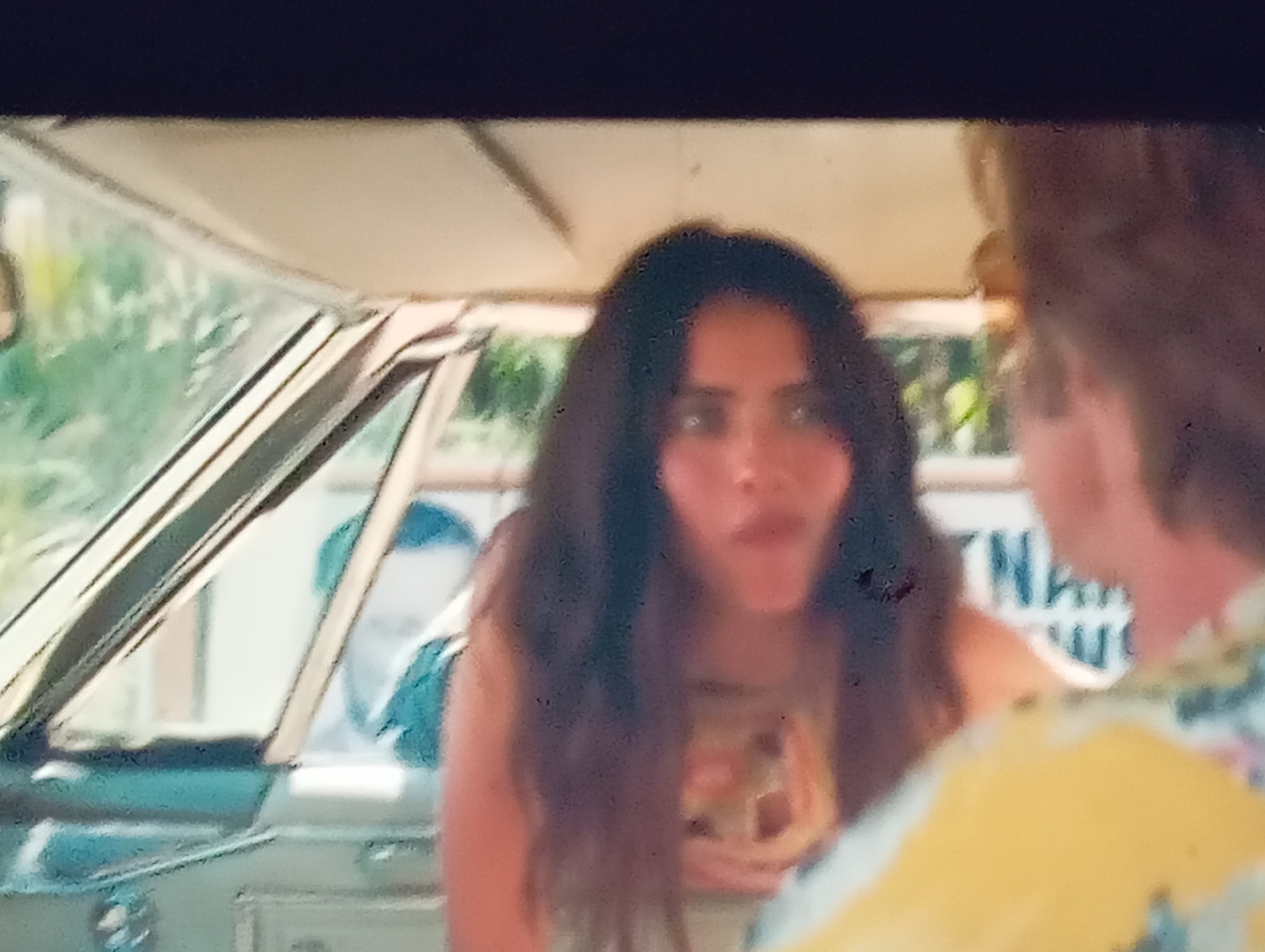 The media file shows a medium shot from inside a car looking out. The primary subject is a young woman with long, dark, wavy hair, wearing a sleeveless top with a colorful, patterned design. She is looking towards the viewer with an open mouth, appearing to be speaking or reacting to something. Her expression is animated.
In the background, through the car windows, lush green foliage is visible, suggesting an outdoor setting, possibly during the day. Another person, a man, is partially visible in the driver's seat, with his back to the camera and wearing a blue shirt. To the right, another person's shoulder and back are visible, wearing a bright yellow and white patterned shirt.
Behind the woman and to the right, there's a white sign with some blurred blue lettering, potentially advertising a business or a location. The interior of the car is visible, with the dashboard and windshield on the left. The lighting suggests it's daytime, possibly sunny, given the brightness and the clear visibility of the outdoor scene. The overall mood seems to be casual and conversational. The location context provided indicates Sofiivska Borshchahivka, Ukraine.