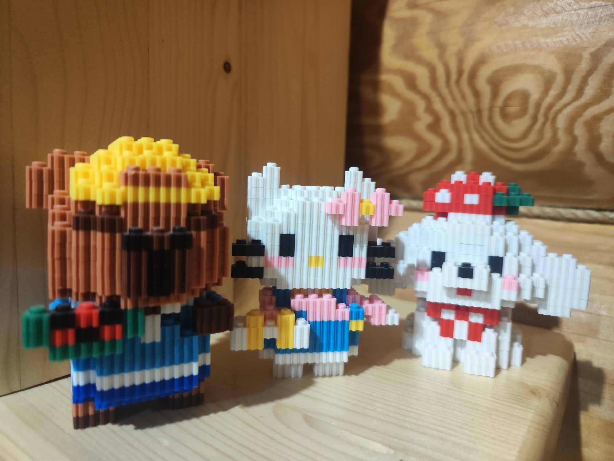 The image shows three pixelated figurines of cartoon characters. From left to right, there is a bear-like character with brown fur, a yellow and brown top, and a blue and white striped outfit. Next to it is a Hello Kitty figurine with a white body, black eyes, a pink bow with a yellow center, and a blue and white outfit. On the right is a white dog-like character with a red and white checkered scarf and a red and white mushroom-shaped hat. The figurines are standing on a light-colored wooden surface, with a light brown wooden background behind them. The location is not discernible from the image.