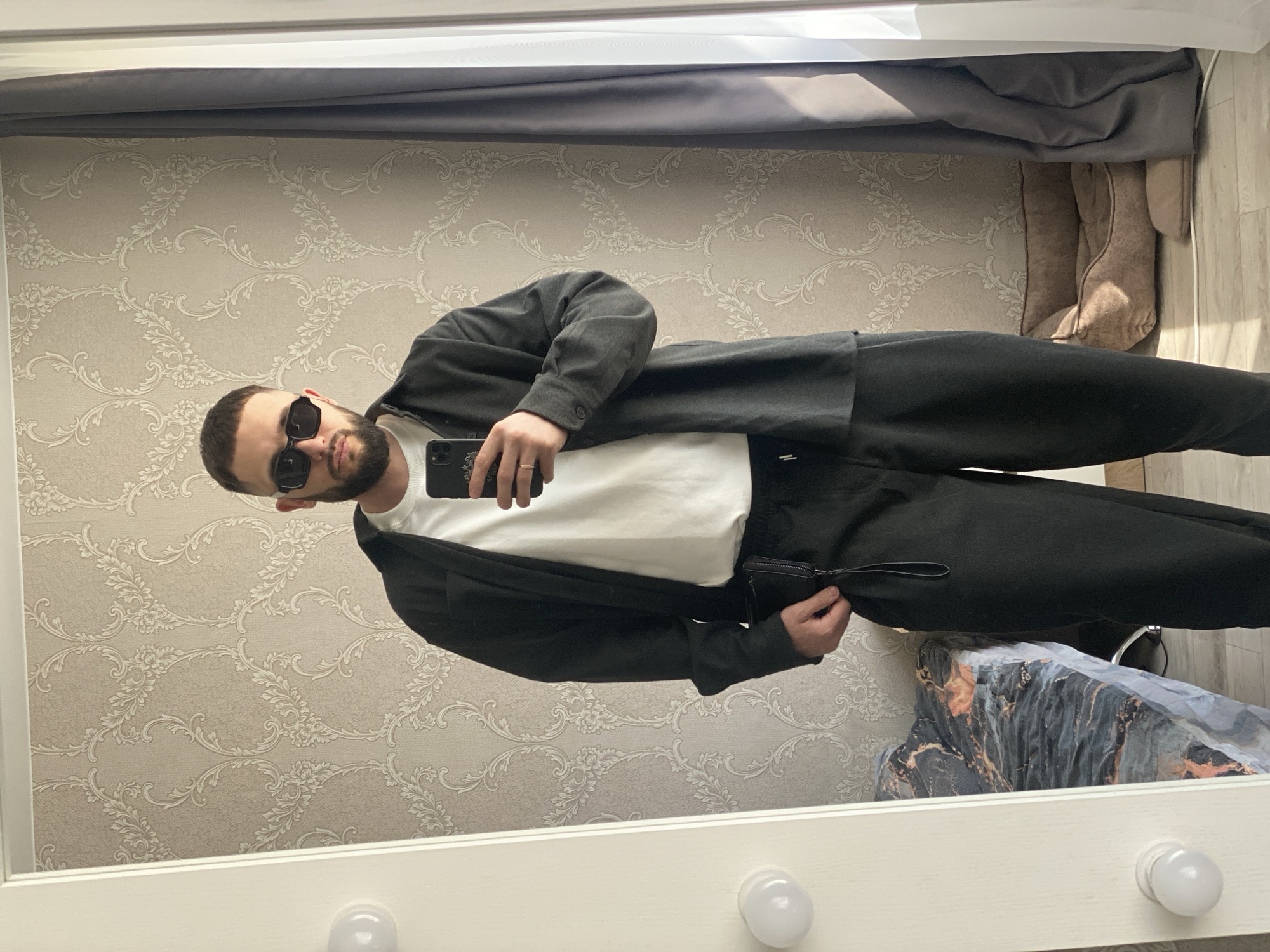 A man with a beard and sunglasses is taking a mirror selfie. He is wearing a white t-shirt and a dark-colored suit or matching set. The mirror has a white frame with round light bulbs along the bottom. The background features patterned wallpaper and a curtain. The man is holding a smartphone, possibly an iPhone, in his left hand. Based on the geotag, the location is likely in Chaikovsky, Russia, although specific details about the precise indoor location are not discernible from the image.