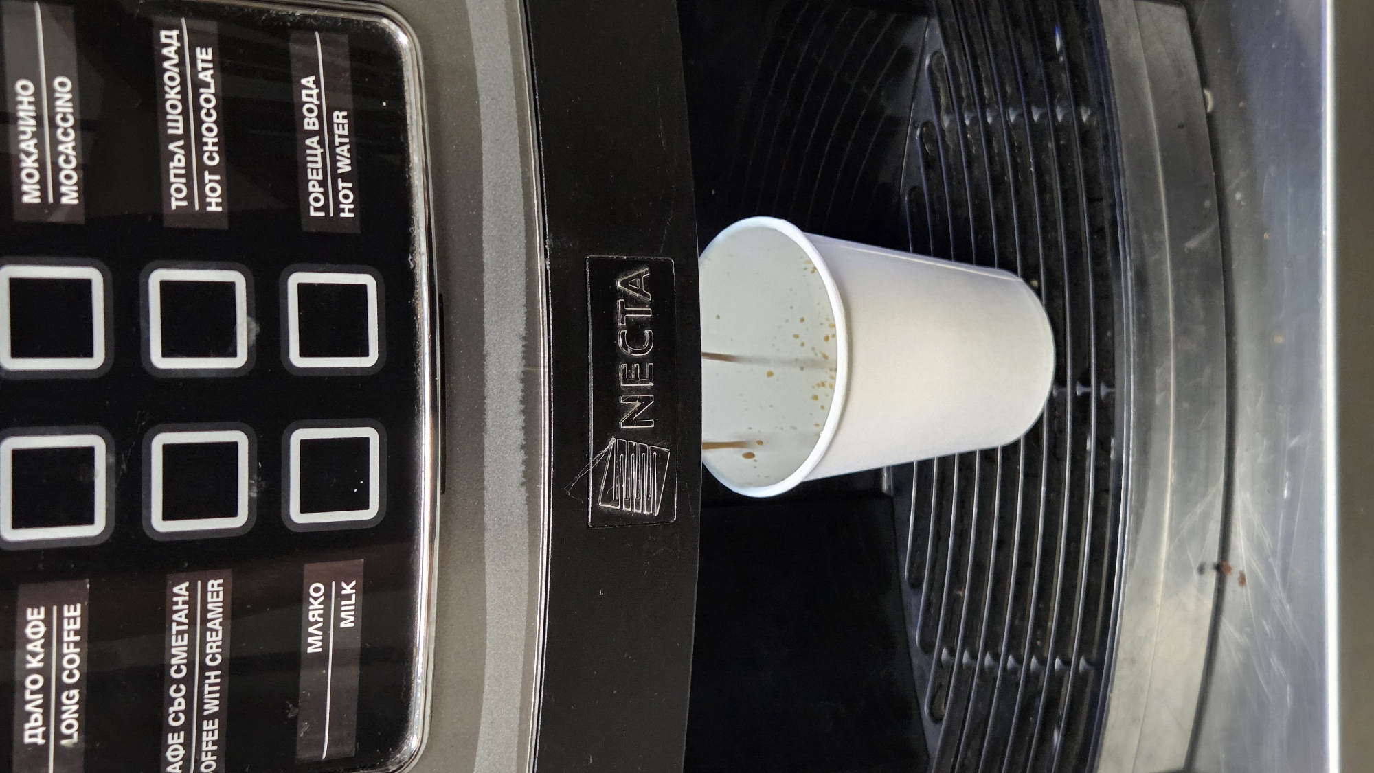 The image shows a close-up of a NECTA coffee machine dispensing coffee into a white paper cup. The machine has a control panel with several buttons, some of which have labels in both Cyrillic and English. The labels include "LONG COFFEE," "COFFEE WITH CREAMER," "MILK," "MOCACCINO," "HOT CHOCOLATE," and "HOT WATER." The coffee is pouring from the machine into the cup, which is positioned on a metal grate. The background is dark, and the overall lighting is somewhat dim. The scene appears to be indoors, likely in a public space such as an office breakroom or a cafe.