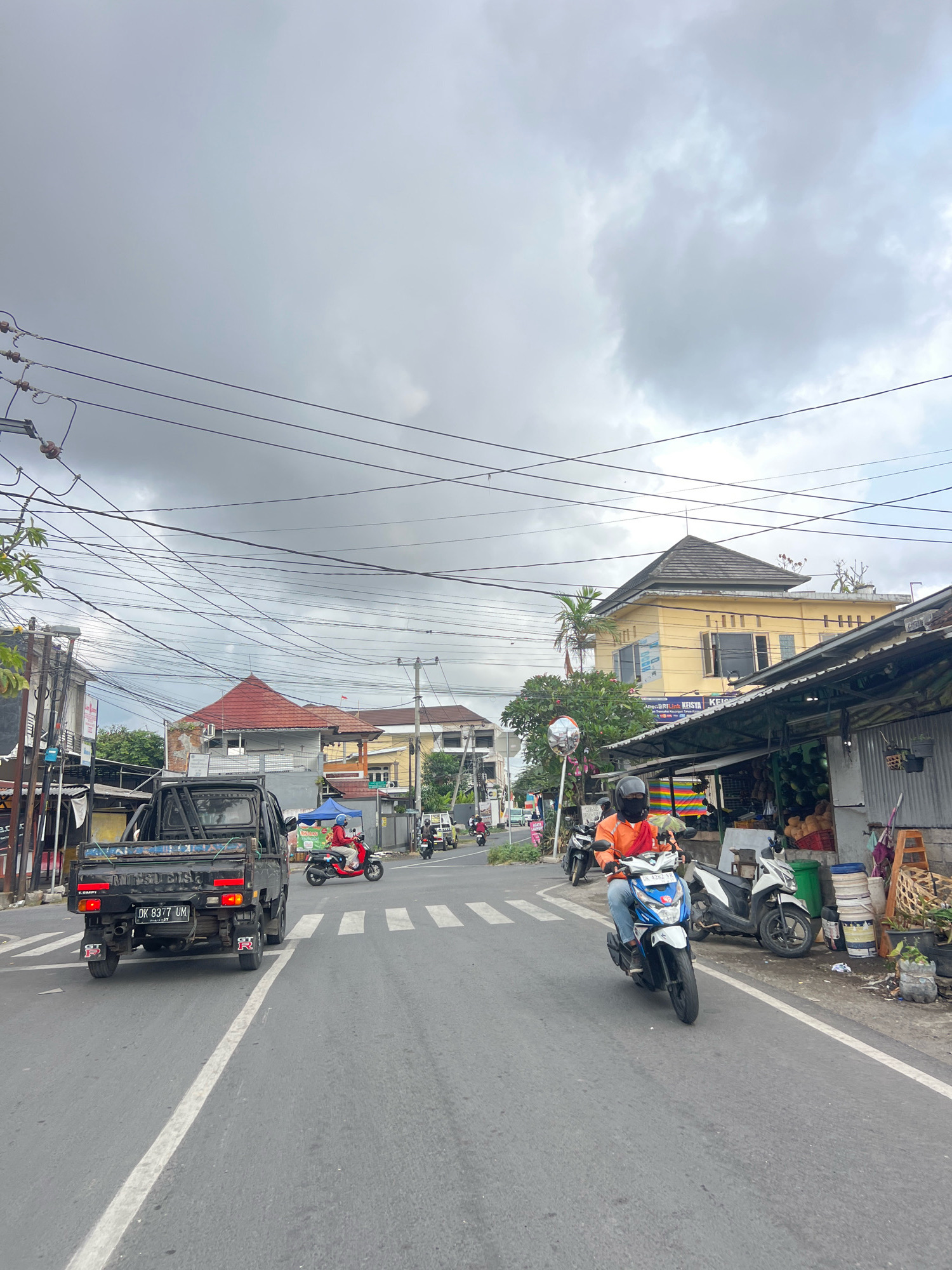 The image shows a street scene in Denpasar, Indonesia, on a cloudy day. A black Mitsubishi pickup truck with a license plate "DK 8377 UM" is driving on the right side of the road. A person wearing a red helmet and blue jacket is riding a red scooter behind the truck. On the right side of the road, a person wearing an orange jacket and blue jeans is riding a white and blue motorcycle. Adjacent to the road, there are several buildings, including a yellow building and a market stall selling fruits and vegetables. Numerous power lines are visible crisscrossing the sky.