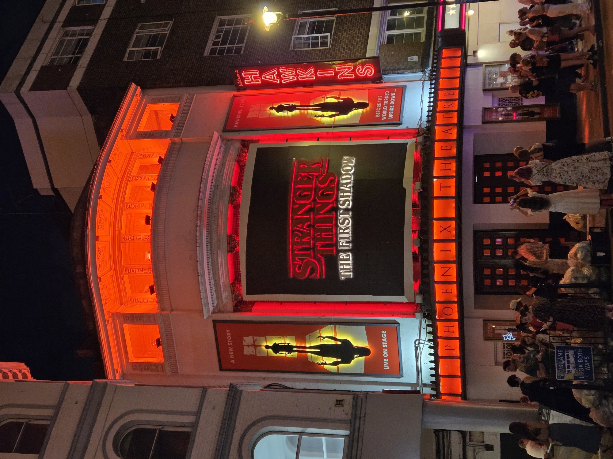Here's a detailed description of the image:
The photograph captures the exterior of the Phoenix Theatre in London, United Kingdom, at night. The theater's facade is illuminated with bright red neon lighting. The marquee displays "STRANGER THINGS THE FIRST SHADOW" in large, glowing red letters. Above and below this central sign are banners advertising the play, featuring a silhouetted figure and taglines like "A NEW STORY" and "LIVE ON STAGE." Above the upper banner is a smaller sign that reads "HAWKINS" in neon, with the phrase "Welcome to HAWKINS" above it. The word "PHOENIX" is vertically spelled out in red neon lights down the side of the building. A crowd of people is gathered outside the theater, some looking at their phones, and some looking at the theatre. Some are dressed in evening attire.
A street sign reading "US LANE LOOK BOTH WAYS" is visible near the lower right corner of the image.