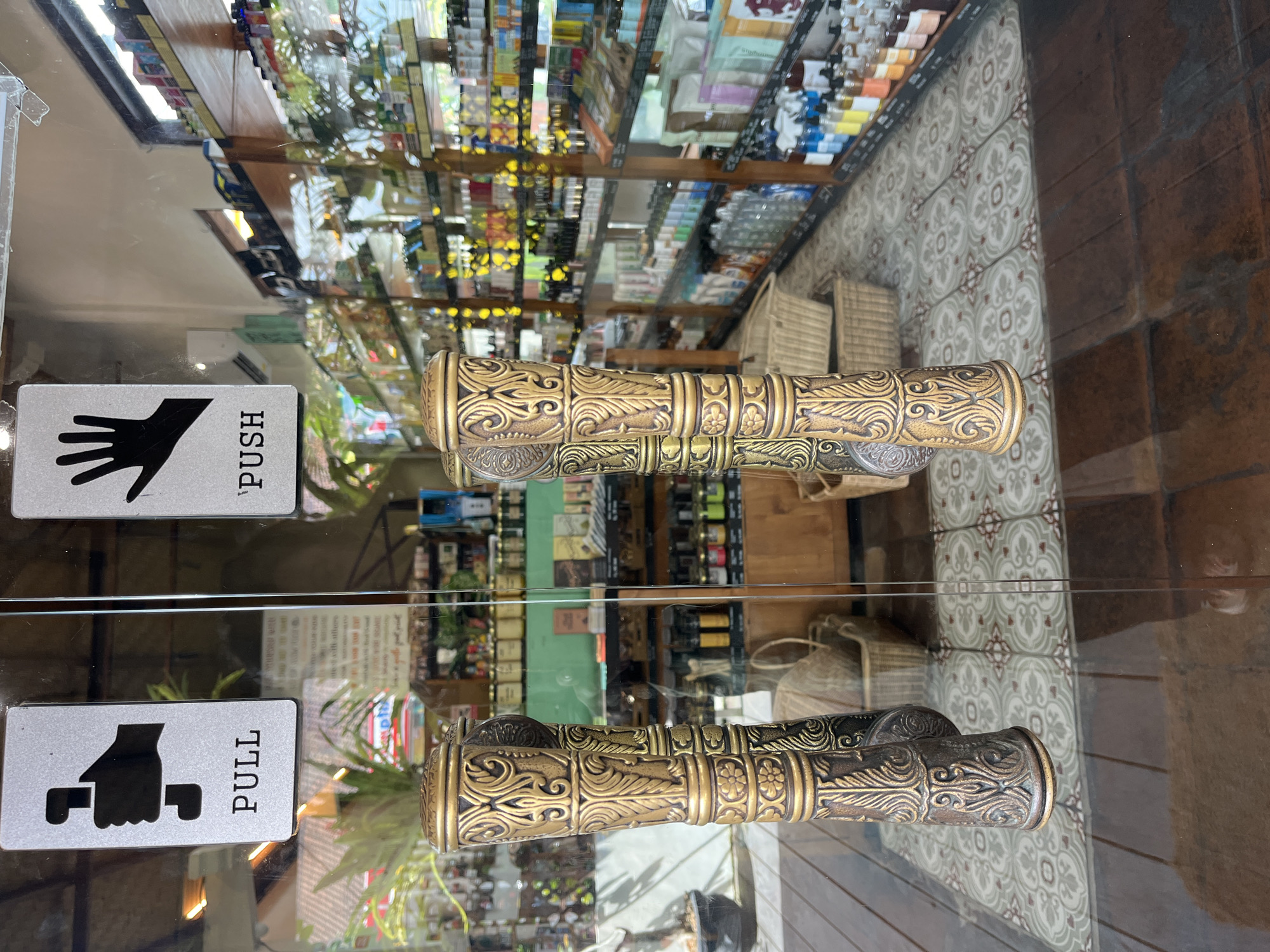 The image shows the entrance to a shop in North Kuta, Indonesia. The entrance has glass doors with elaborate gold and bronze colored handles. Above each door handle, there are signs indicating "PULL" and "PUSH". The shop interior is visible through the glass doors, revealing shelves stocked with various products, including bottles, jars, and boxes. Wicker baskets are also visible on the floor inside. The floor in front of the doors is tiled with a patterned design, and beyond that, wooden floorboards are present. The overall impression is of a well-stocked retail establishment with a decorative aesthetic.
