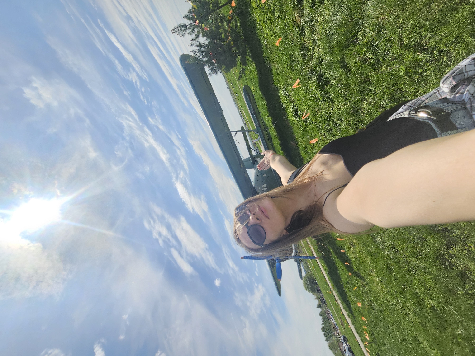 A medium-shot, slightly angled image shows a woman in sunglasses taking a selfie on a sunny day, with a biplane in the background. The background is a grassy field with a treeline and some distant structures. The location is possibly the Izhevsk Aviation Museum in Russia. The woman is wearing a black tank top and denim shorts with a plaid shirt tied around her waist. She is positioned on the right side of the frame, her right arm outstretched to take the selfie. The biplane is positioned behind her, spanning the center of the frame, with its wings perpendicular to the image plane. The bright sky is visible in the upper left corner, while the sun's rays streak across the sky. A line of orange flags lines the horizon near some cars parked nearby.