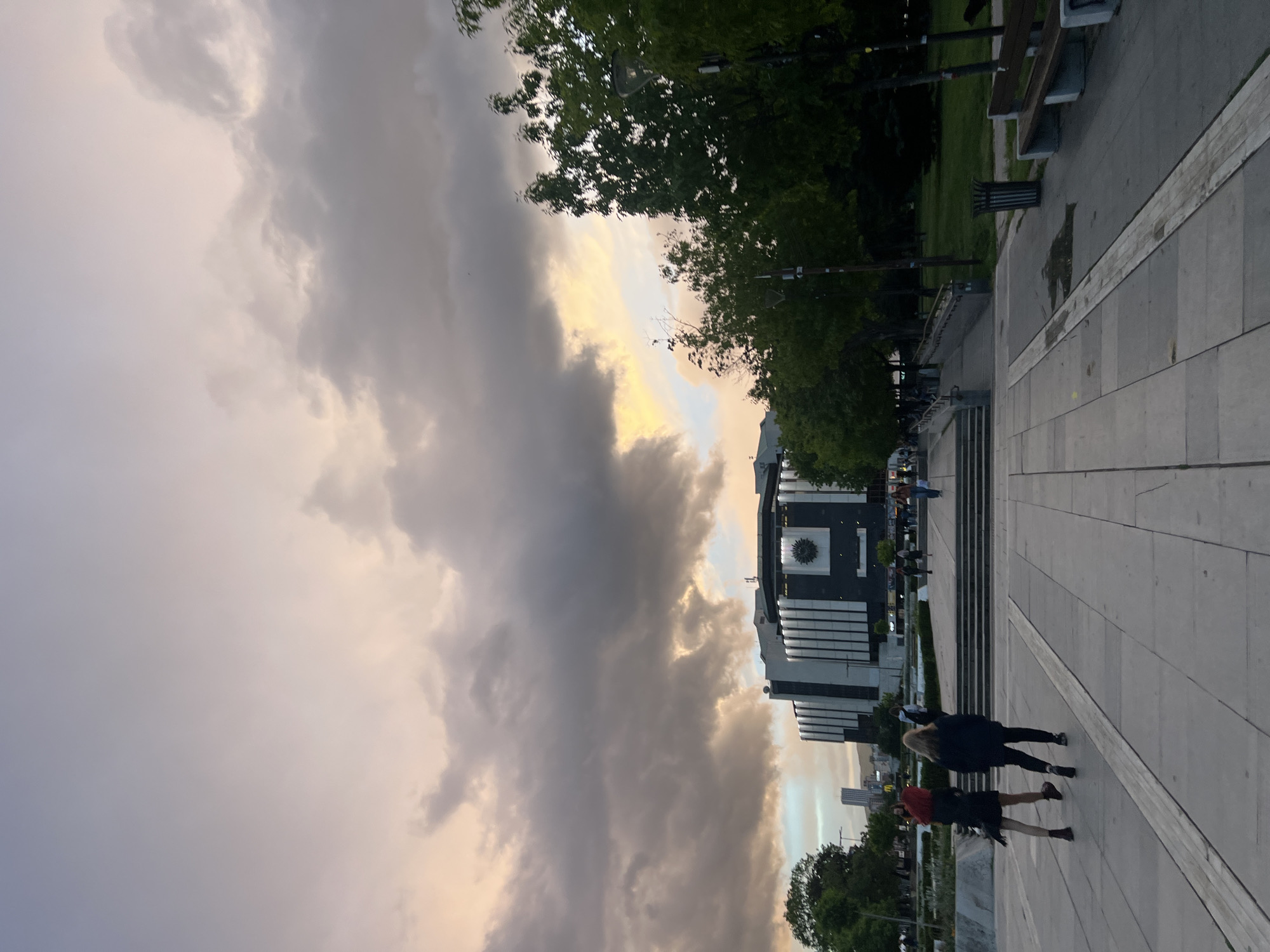 The image shows a scene in Sofia, Bulgaria, featuring the National Palace of Culture (NDK) under a dramatic sky. The sky is filled with dark, heavy clouds with hints of golden light peeking through, suggesting either sunset or the moments before a storm. Below the clouds is a large building, the NDK, notable for its modern architectural style with vertical lines and a large clock face. In the foreground, there is a paved area with a few people walking. One pair is closer to the viewer and appears to be a person with red hair, dressed in a dark outfit, walking alongside another person with long blonde hair, also dressed in a dark outfit. The paved area is bordered by trees and park benches, which are lined along a grassy area on the side. The composition is wide, capturing both the imposing building and the figures traversing the plaza.