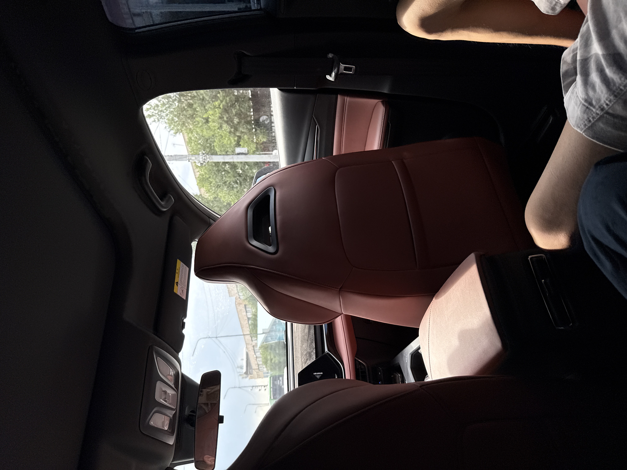 The image depicts the interior of a modern car, viewed from the rear passenger seats. The seats are upholstered in a rich, dark red leather, with a sporty design featuring prominent bolsters and cutouts. In the foreground, the back of the driver's seat and the passenger seat are visible, along with a portion of the center console with a grey, textured surface and a metallic accent. The dashboard and a large infotainment screen are visible between the front seats.
The ambient light suggests it is daytime, with sunlight streaming in through the windows, casting shadows within the car. The exterior view through the windows reveals a street scene with trees, buildings, and power lines, characteristic of an urban environment. A green bus is visible on the road outside, and there are parked cars and a light pole.
The image focuses on the car's interior, with a person's legs visible in the lower right corner, wearing shorts. The overall impression is one of a well-appointed vehicle in a city setting. Given the provided context of Almaty, Kazakhstan, the street scene, building architecture, and vegetation are consistent with an urban area within that city.