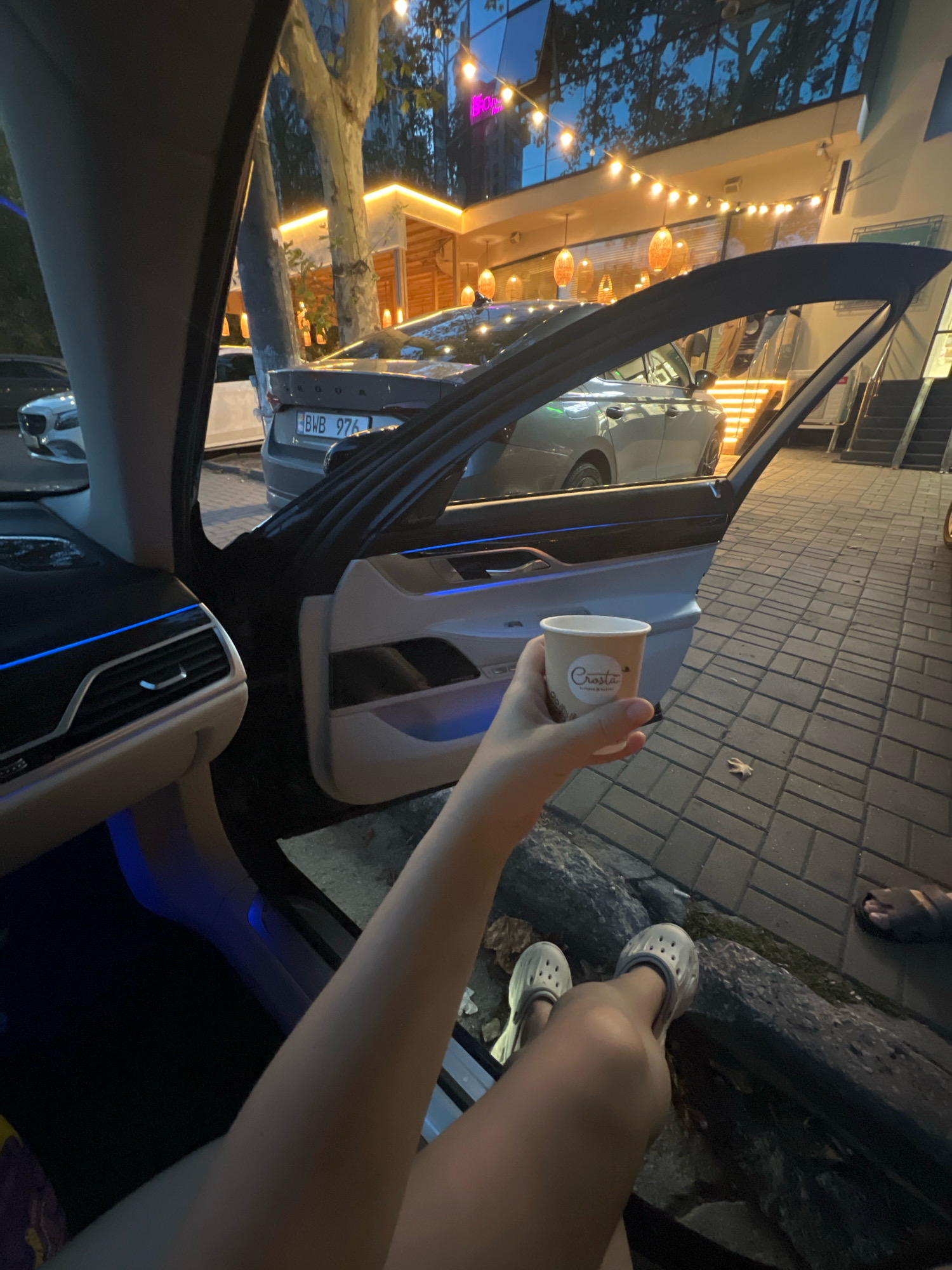 In this medium shot, taken from inside a car with the driver's side door open, a person's arm and legs are visible. The person is holding a paper coffee cup with a logo that reads "Crosta". They are wearing white Crocs. The car's interior features ambient blue lighting. Outside the car, a paved sidewalk is visible, along with parked cars and a building with illuminated string lights. The scene appears to be a casual stop in an urban setting at dusk or night.