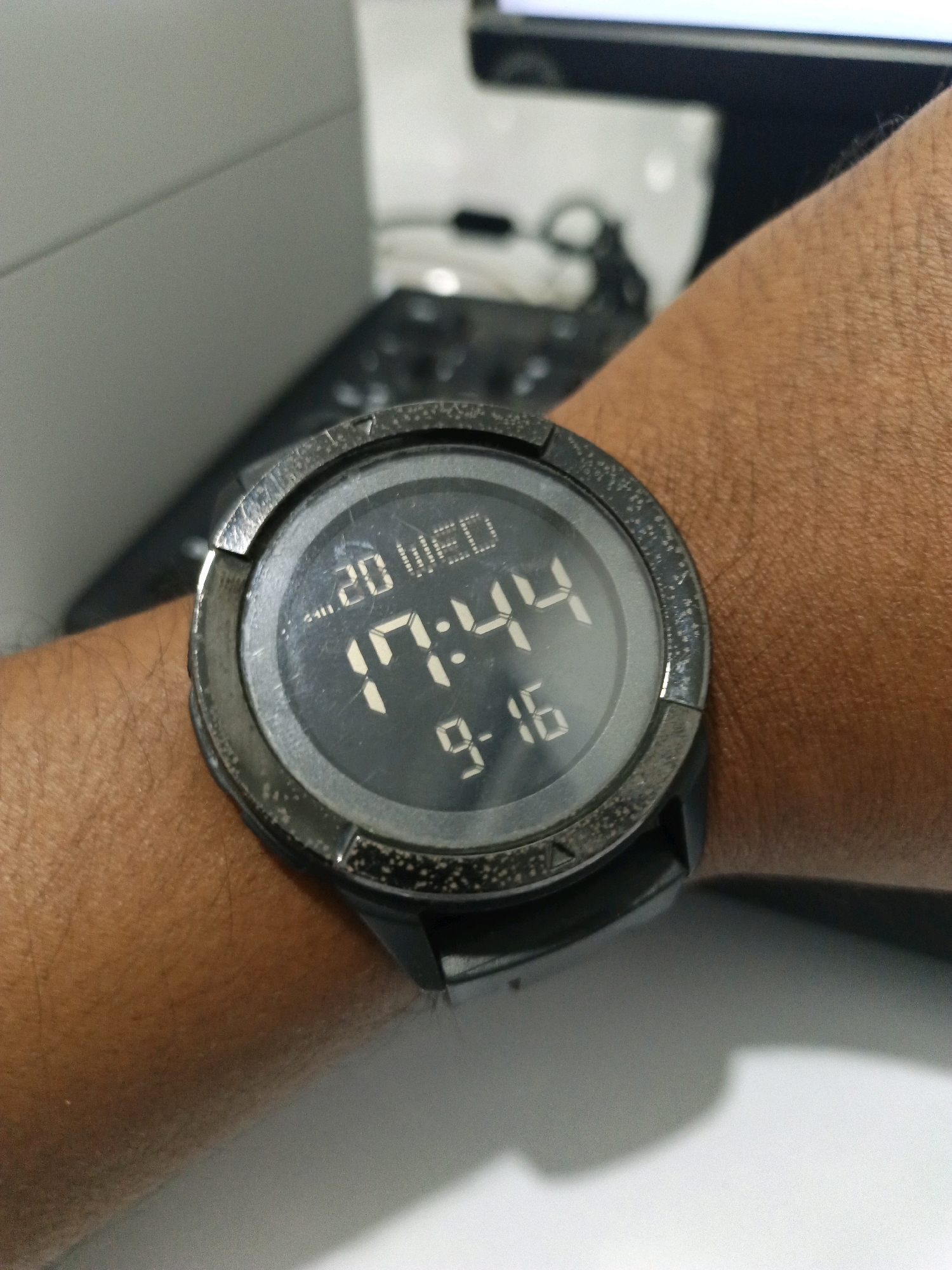 A close-up shot shows a person's wrist wearing a digital watch. The watch face displays the date "20 WED" and the time "17:44" with the minutes "9-16" below. The watch has a dark bezel and a black strap. The wrist appears to be a light to medium skin tone with visible hair. In the background, a keyboard is partially visible, suggesting the person might be at a desk. The lighting is adequate, highlighting the details of the watch and wrist. The background is blurred, with a gray surface and a hint of a monitor. The image appears to be taken indoors, possibly in an office or study environment. The location is not specified within the image itself.