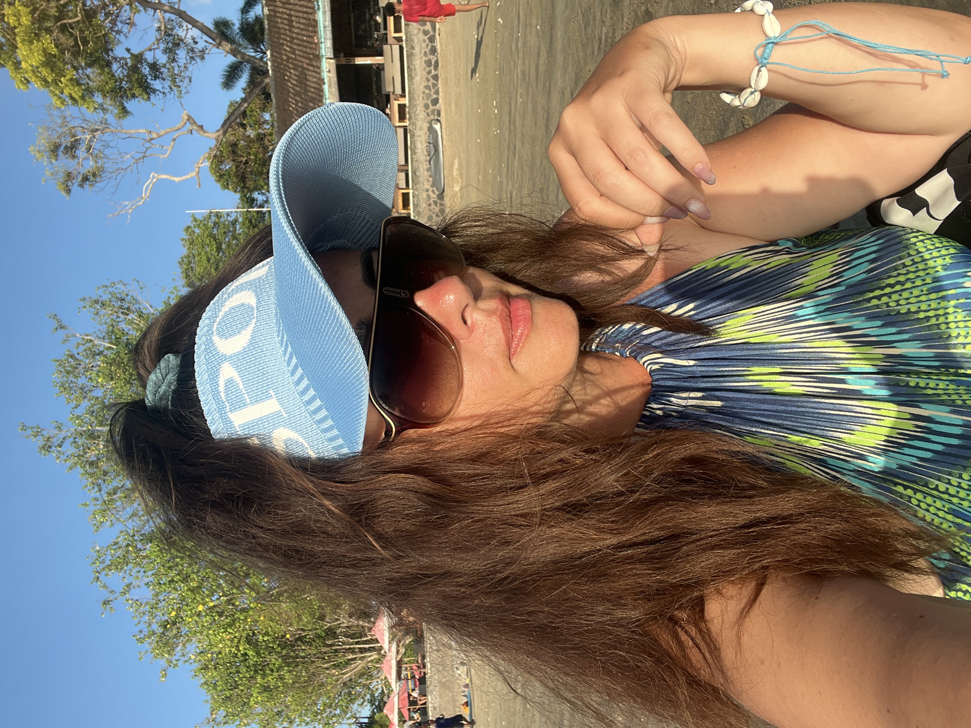 An outdoor, medium-shot selfie shows a woman with long, brown hair and a blue visor with white lettering. She is wearing large, brown sunglasses and a blue, green, and white patterned halter top. She is on a beach, with a person in a red shirt walking in the background. Trees and buildings are visible behind the woman, suggesting the location is in Kuta Selatan, Indonesia. A bracelet made of shells is visible on her wrist.