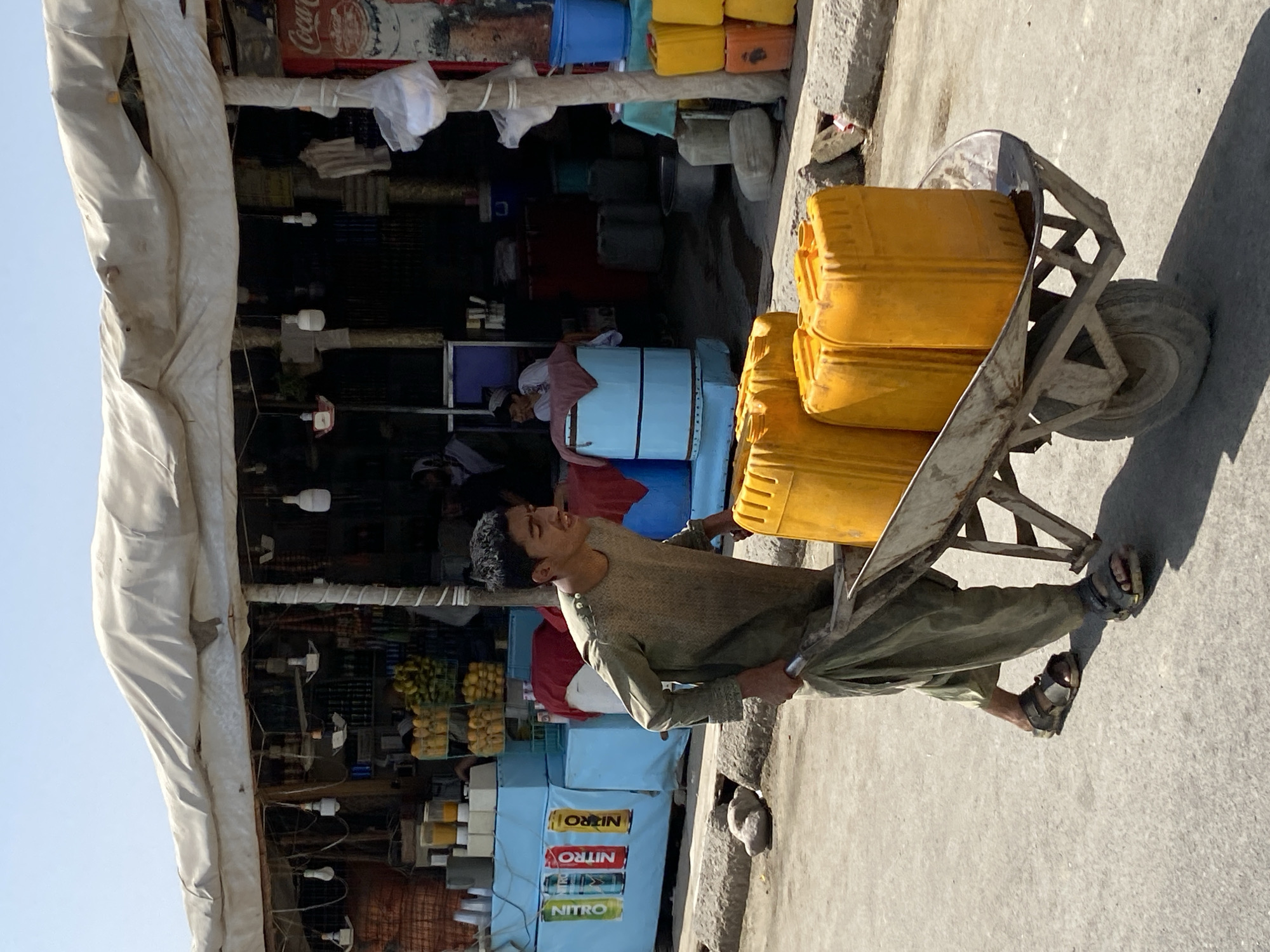 In the city of Kandahar, Afghanistan, a young man is pushing a wheelbarrow filled with yellow plastic containers along a dusty street. The man, dressed in a loose-fitting olive green garment and sandals, walks with his head down, focused on his task. The wheelbarrow is loaded with at least three large, rectangular yellow containers, possibly filled with water or another liquid. Behind him, a makeshift market stall is visible, covered by a white tarpaulin. The stall displays various goods, including crates of bottled drinks with colorful labels such as "NITRO," stacks of fruit, and other items. Several electric lights hang from the stall's structure, illuminating the products. In the background, more stalls and structures are present, suggesting a busy marketplace. The overall scene depicts a typical day in a bustling Afghan city, with commerce and daily life in full swing under a clear, bright sky.