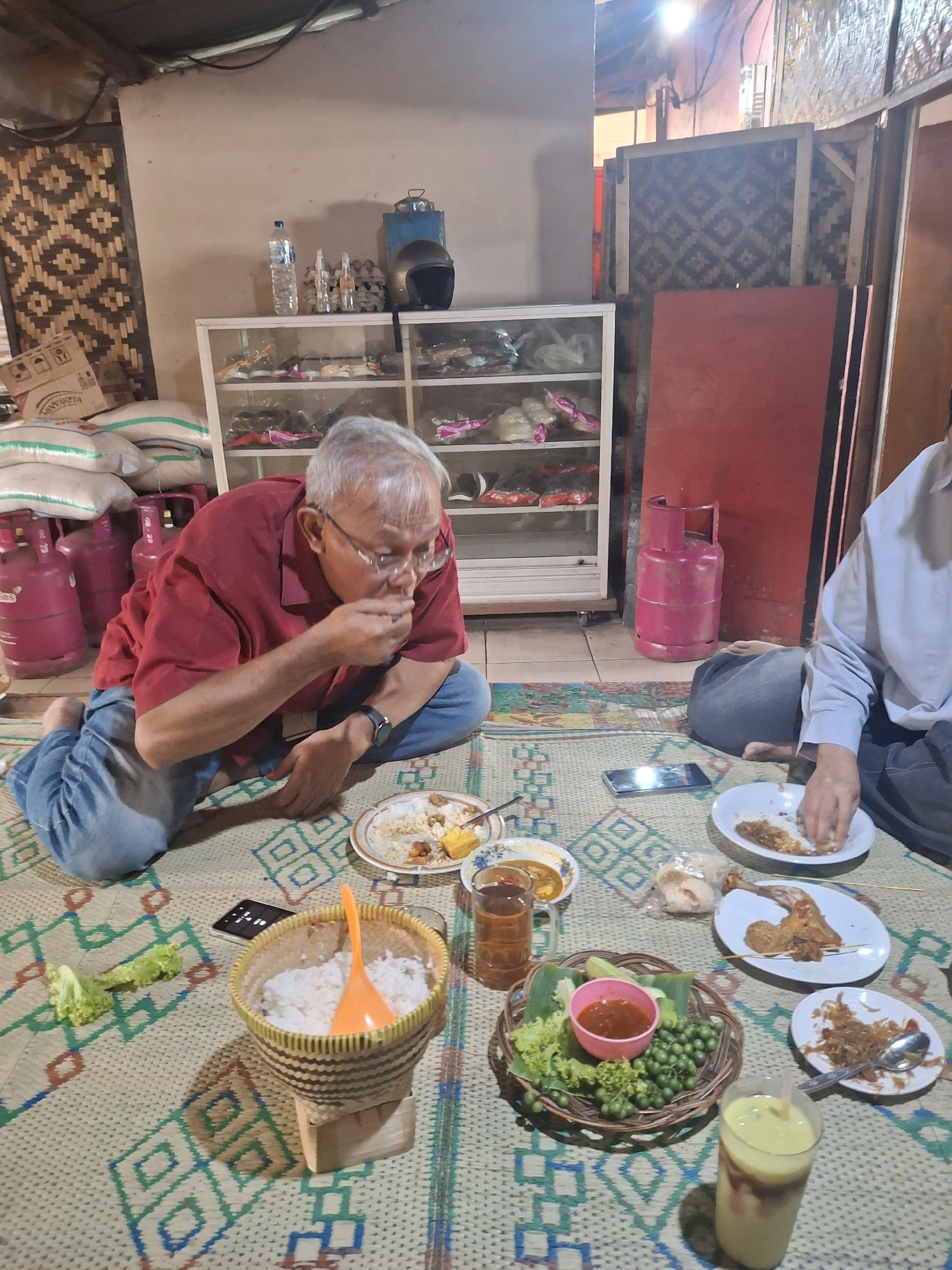 In Cibogo, Indonesia, a man in a red shirt and blue jeans is sitting on a woven mat, eating rice from a plate. He is wearing glasses and has gray hair. The mat is spread on the floor of a room with a wooden wall behind him, decorated with a geometric pattern. To his left, there are several pink gas cylinders and bags of rice. A white cabinet filled with various items stands against the wall. In front of the man, a variety of dishes are laid out on the mat, including rice, chicken, vegetables, and sauce. Another person, wearing a light blue shirt, is partially visible in the foreground, also eating. The scene appears to be a casual meal in a home or a modest establishment.
