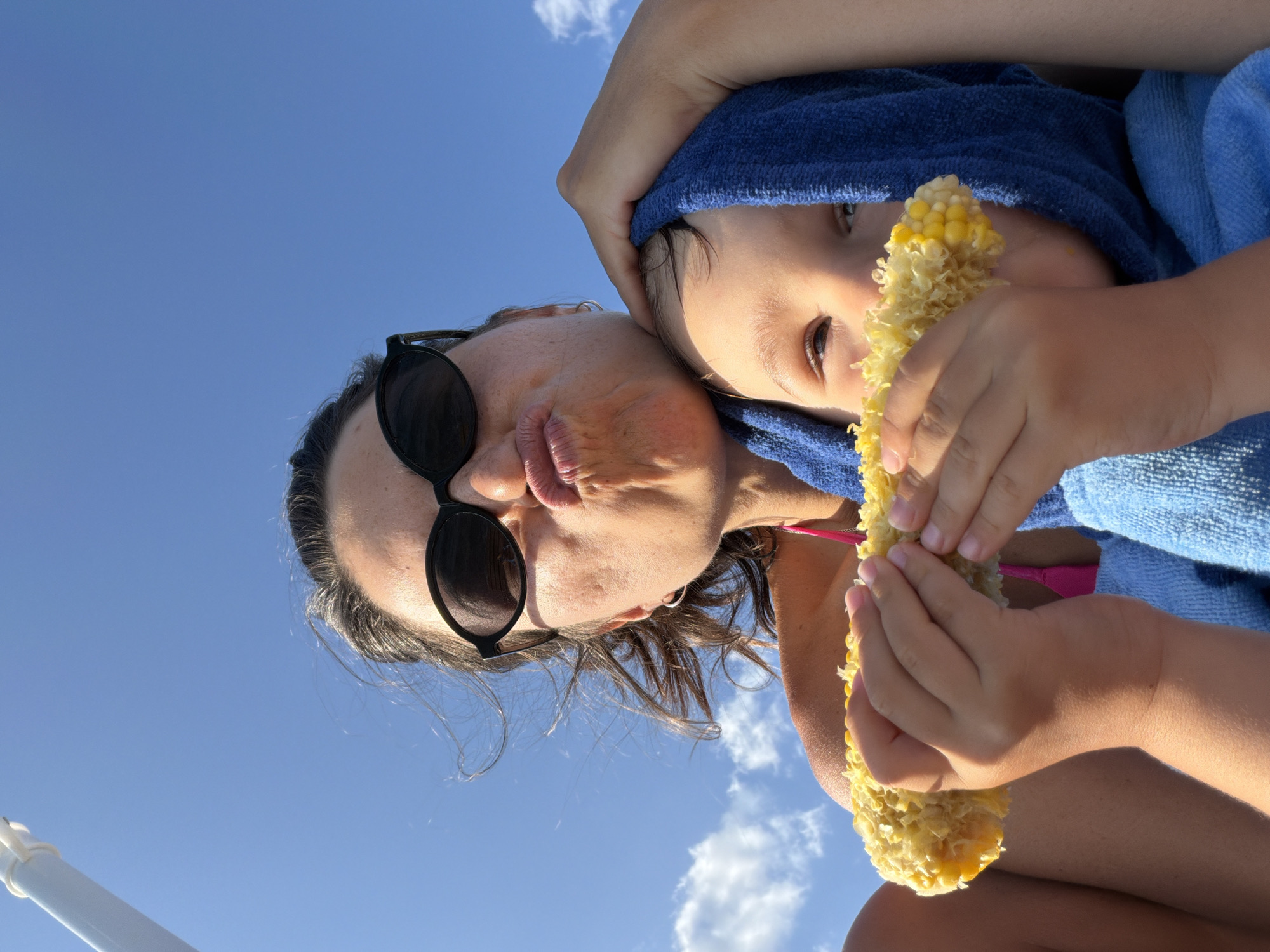 The image shows a woman and a child in Budva, Montenegro, against a bright blue sky with scattered clouds. The woman, positioned in the upper half of the frame, is wearing sunglasses and making a puckered face. She has her arm around the child. The child, in the lower half of the frame, is wrapped in a blue towel and is holding a cob of corn with both hands, taking a bite. The corn cob is partially eaten. The overall scene suggests a casual, outdoor setting, likely on a sunny day.