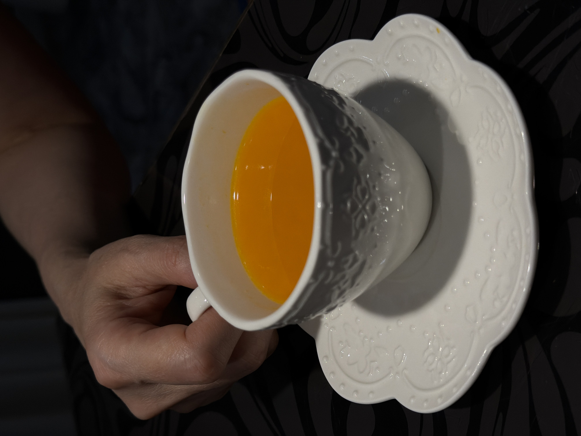 A person's left hand is holding a white teacup filled with an orange liquid, presumably tea or juice. The cup sits on a white saucer with a decorative edge. The background is a dark, patterned surface, possibly a tablecloth or table. The location is unspecified and cannot be determined from the image alone.