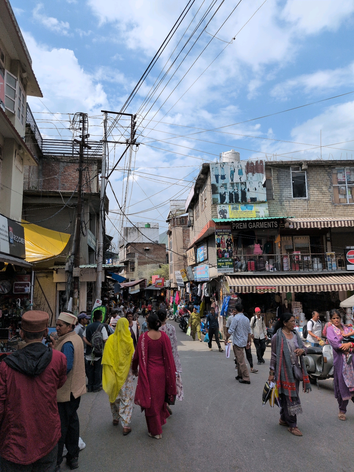 The image depicts a bustling street scene in Bhuntar, India, with a focus on a crowded pedestrian thoroughfare. Numerous people are walking in both directions, some singly and others in small groups. They are dressed in a variety of casual and traditional Indian attire, including salwar kameez and kurtas. Many are carrying shopping bags, suggesting a market or commercial area.
The street is lined with buildings on both sides, featuring shops and businesses with awnings and signage, some of which are in English and Hindi. Electrical wires crisscross overhead, creating a dense network against a partly cloudy blue sky. Several utility poles are visible, supporting the extensive wiring.
In the foreground, men and women are seen walking along the asphalt road. On the left side, a man in a maroon kurta and a brown turban is walking away from the camera. To his right, another man in a beige vest and a white cap is facing him. A woman in a bright yellow garment is walking away from the camera, followed by another woman in a dark pink kurta. Further down the street, more individuals are moving through the crowd.
On the right side of the street, a silver scooter is parked, and a woman in a purple salwar kameez is standing beside it, holding shopping bags. Another woman in a similar outfit is walking away from the scooter. Shops display various goods, including clothing and textiles. One building prominently features a large advertisement for "PREM GARMENTS" and "SCHOOL DRESSES," with images of children in school uniforms. The overall impression is one of vibrant daily life and commerce in a busy Indian town.