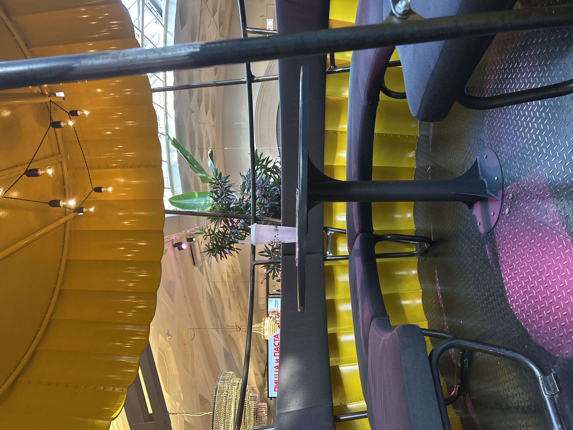 A circular booth seating area with dark gray upholstered benches is illuminated by a bright yellow, corrugated wall. A black circular table with a single pedestal base stands in the center of the booth. A pink and white striped cup with a straw sits on the table. Above the booth, a large yellow umbrella-like structure is adorned with a string of exposed lightbulbs. In the background, a lush green plant is visible, and further beyond, a large crystal chandelier hangs from the ceiling of what appears to be a shopping mall or public indoor space. The floor is covered in diamond plate metal with pink circular accents.