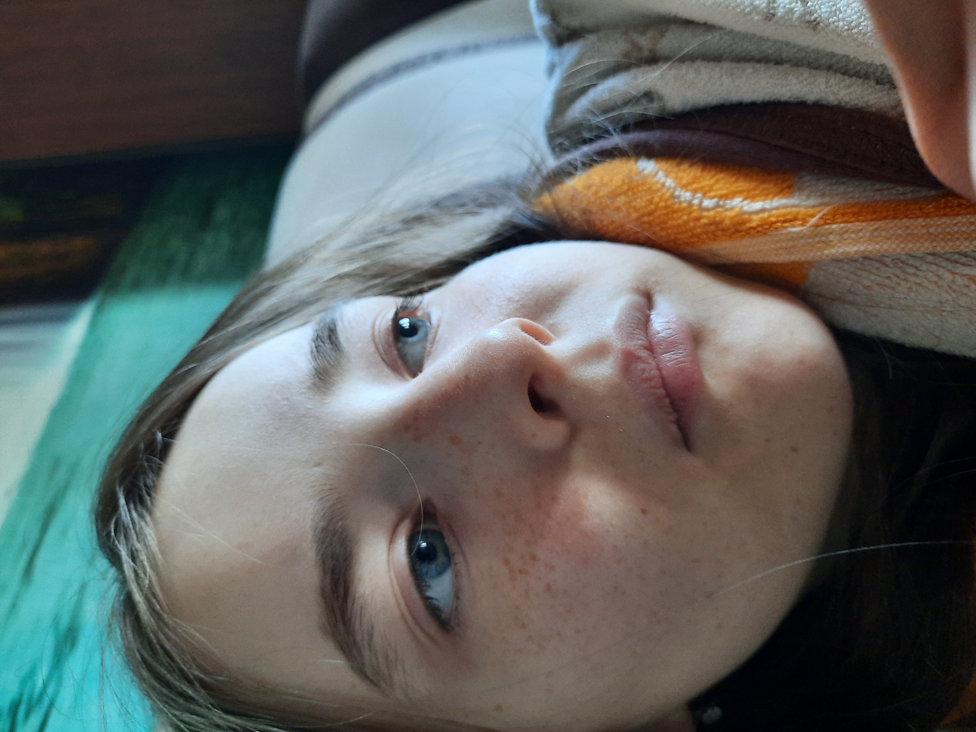A close-up, tilted portrait shows a young woman with fair skin and blue eyes. She has freckles on her face and light brown hair. She is lying down, with a striped towel near her head. Parts of the image contain a turquoise surface. I have no information available to confirm if this image was taken in the city of Архангельское, Russia.