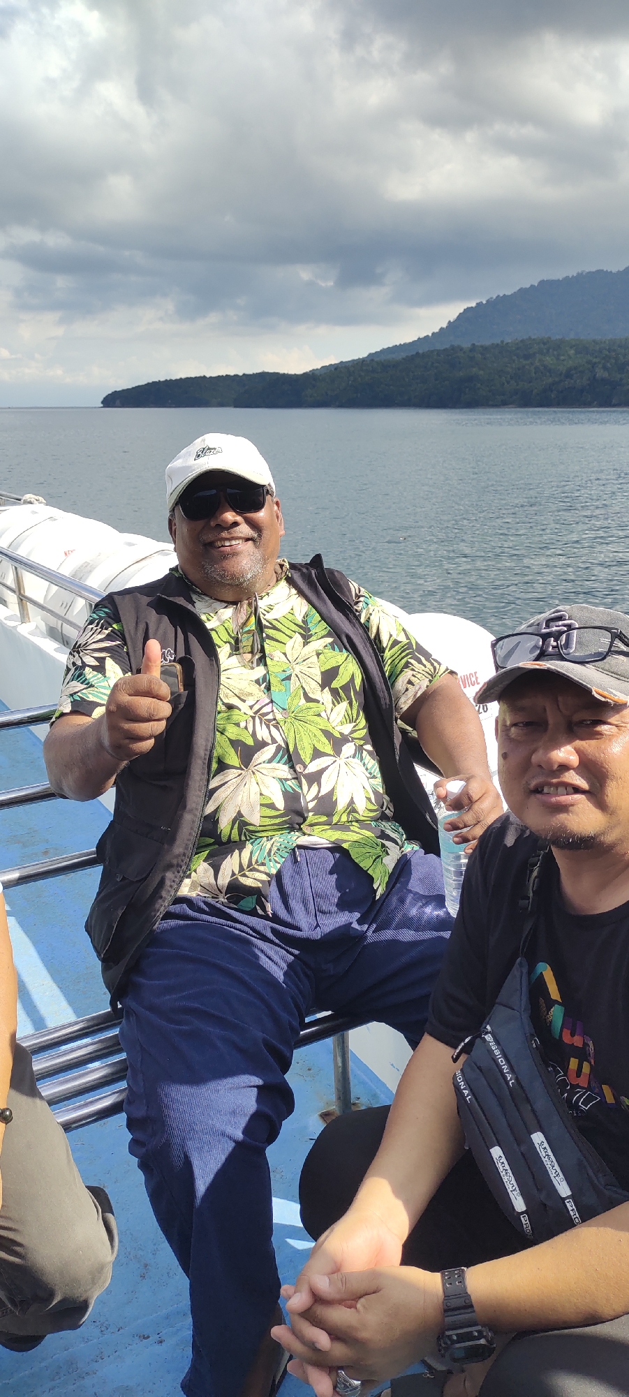 Two men are seated on a boat with a body of water and a forested island in the background. The man on the left is wearing a white cap, sunglasses, a black vest over a tropical-print shirt, and blue pants. He is giving a thumbs-up gesture with his right hand and holding a bottle of water in his left. The man on the right is wearing a black cap, sunglasses, and a black t-shirt. He has a crossbody bag with text printed on it. The boat appears to be on the waters surrounding Sabang, Indonesia, an island city known for its natural beauty and diving spots. The sky is overcast with clouds.