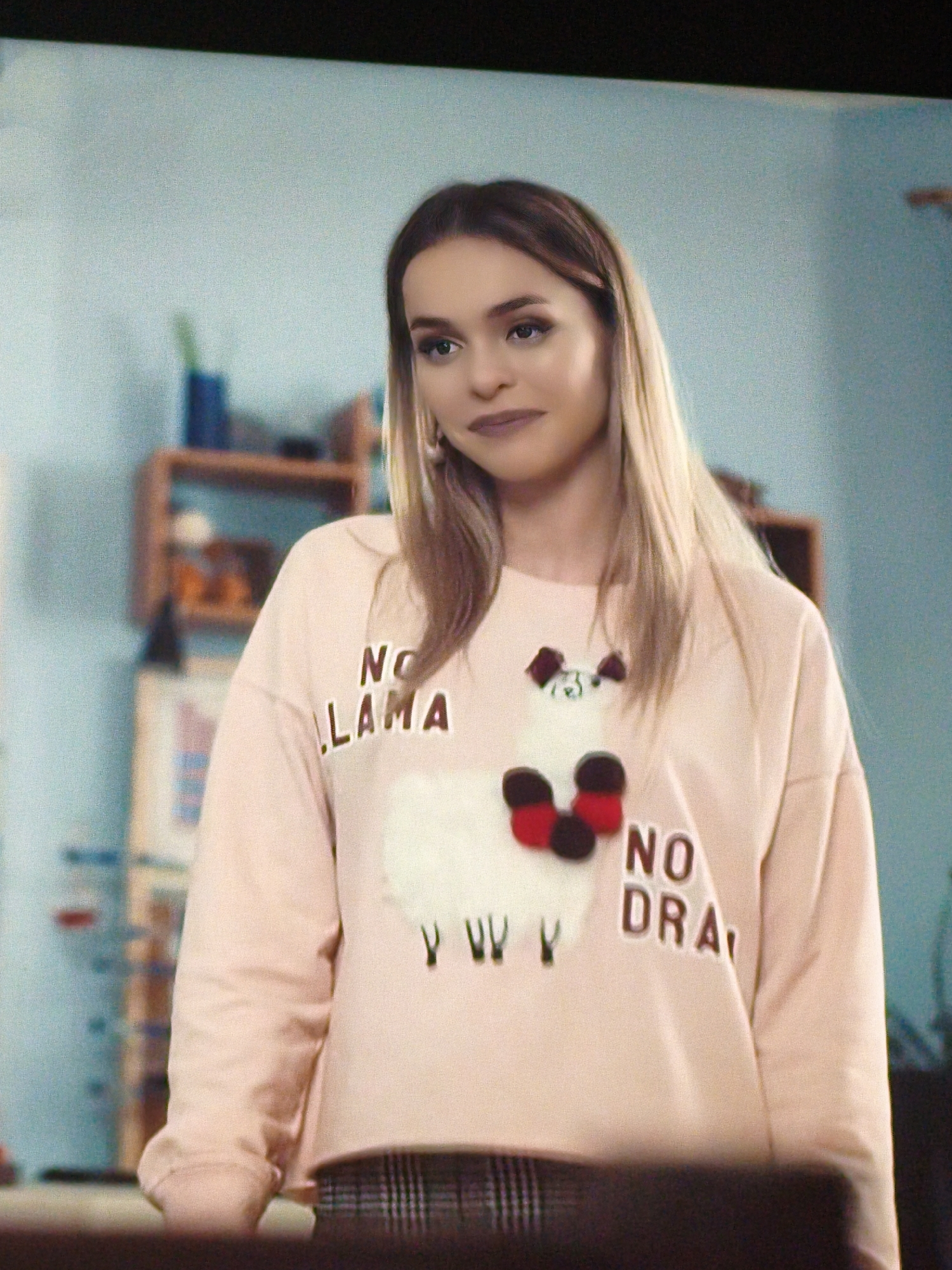 The image shows a young woman with blonde hair, wearing a light pink sweatshirt with a graphic of a llama and the text "NO LAMA" and "NO DRA". She is standing indoors, with a pale blue wall and shelves filled with various objects in the background. The environment appears to be a room in a home or an office. There is no indication of the city of Vladivostok, Russia, in the image.