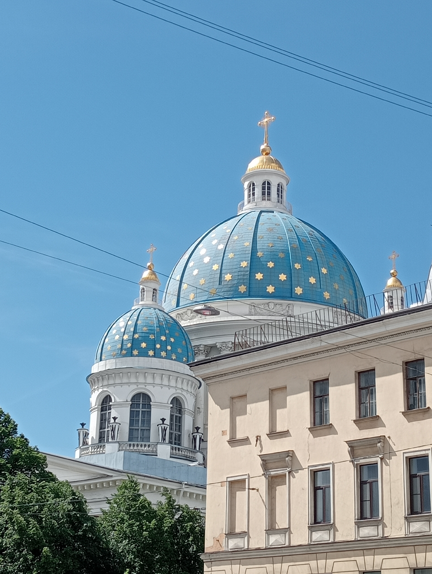 The image captures an upward view of the Trinity Cathedral in Saint Petersburg, Russia. The cathedral features striking blue domes adorned with golden stars and topped with golden crosses. Two main domes are visible, with the larger one dominating the foreground and a smaller one to its left. A portion of a pale yellow building with numerous windows is situated in the lower right quadrant of the image, partially obscuring the cathedral's lower structures. Lush green trees are visible in the lower left, adding a natural element to the urban landscape. Overhead, several dark power lines traverse the clear blue sky, indicating the presence of infrastructure. The scene is bathed in bright daylight, casting distinct shadows and highlighting the architectural details of the cathedral and the surrounding buildings.