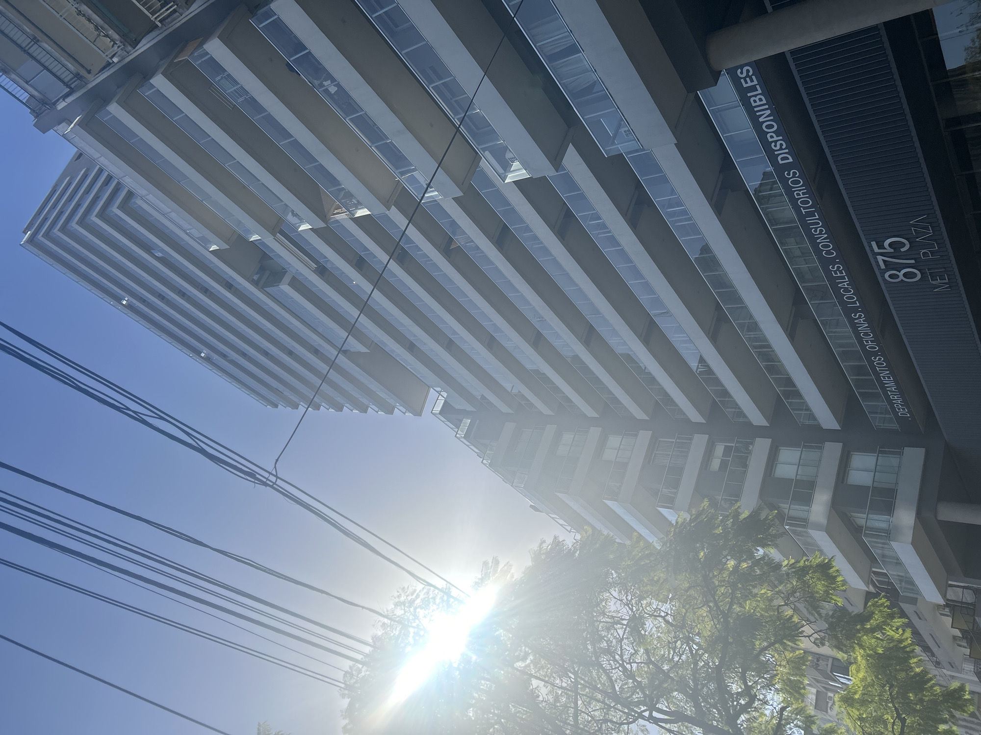 The image shows a modern apartment building in Buenos Aires, Argentina, with the street number "875" and "MET PLAZA" visible on its facade, indicating its location. The building features a repeating pattern of balconies with glass railings. Overhead, several utility wires stretch across the frame against a clear blue sky. The sun is bright and positioned to the left, casting a lens flare and illuminating the scene. To the left of the building, a tree with green foliage is visible. The perspective is from a low angle, looking upwards at the structure.