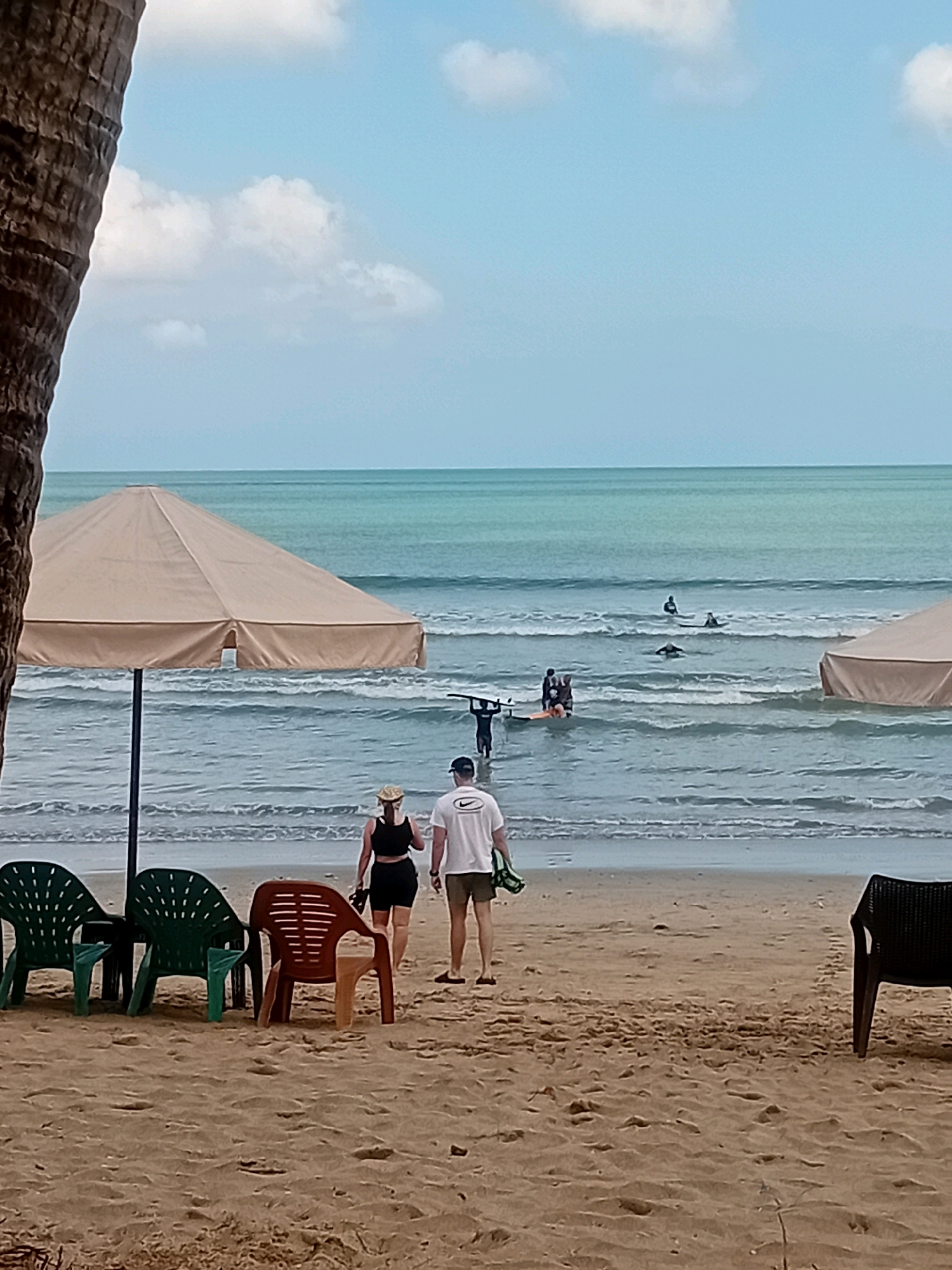This image captures a scene on a sandy beach in Kuta, Indonesia. In the foreground, several plastic chairs, two green and one orange, are arranged on the sand. To the left, the rough bark of a palm tree trunk is visible. Further back, two people, a woman in a black top and shorts and a man in a white t-shirt and shorts, walk away from the viewer towards the ocean. A large, beige beach umbrella stands between them and the water.
In the shallow waters of the ocean, several individuals are engaged in activities related to surfing. Some are standing on surfboards, while others are in the water with their boards. The ocean is a vibrant turquoise color, with gentle waves rolling towards the shore. The sky above is a light blue with scattered white clouds. The overall environment suggests a relaxed beach day.