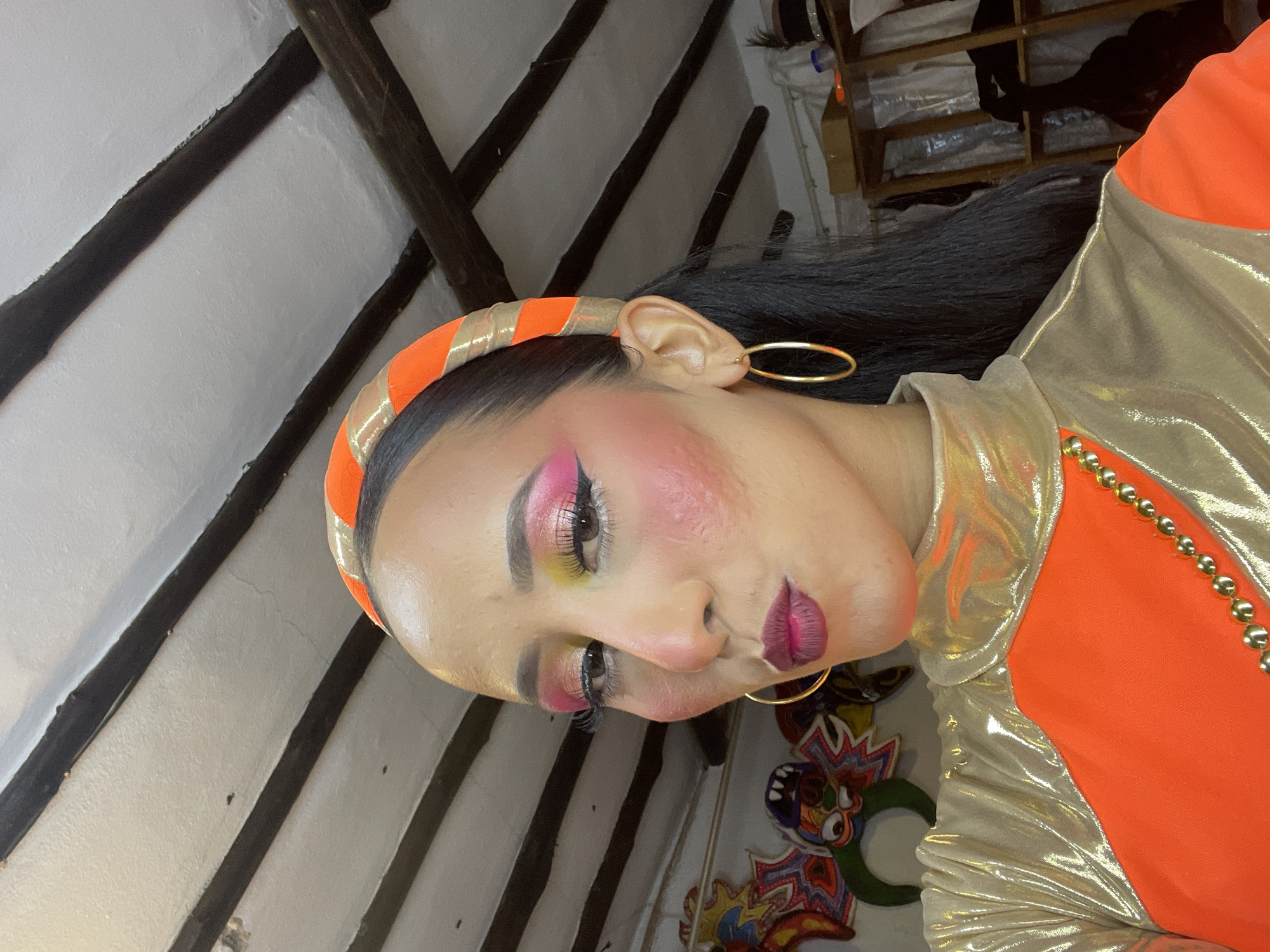 A close-up, medium shot shows a woman with dramatic makeup and a vibrant orange and gold outfit. She is looking at the camera with her mouth slightly pursed and her head tilted. Her eyes are lined with black eyeliner and adorned with false eyelashes, and her eyelids feature a bold gradient of pink and yellow eyeshadow. She has prominent blush applied to her cheeks and dark lipstick. A headband with orange and gold stripes adorns her head, and a dark ponytail extends down her back. She wears large gold hoop earrings.
The woman is wearing a high-necked, shiny gold top layered over an orange garment. A beaded gold necklace is visible across the orange fabric. In the background, a white ceiling with dark wooden beams is visible. To the left, colorful carnival masks or decorations are partially visible. To the right, shelves with various items, including boxes and what appear to be garments, are present. The lighting is bright, casting some shadows. The scene appears to be indoors, possibly a dressing room or studio space, but there is no specific indication of its location in Porlamar, Venezuela.