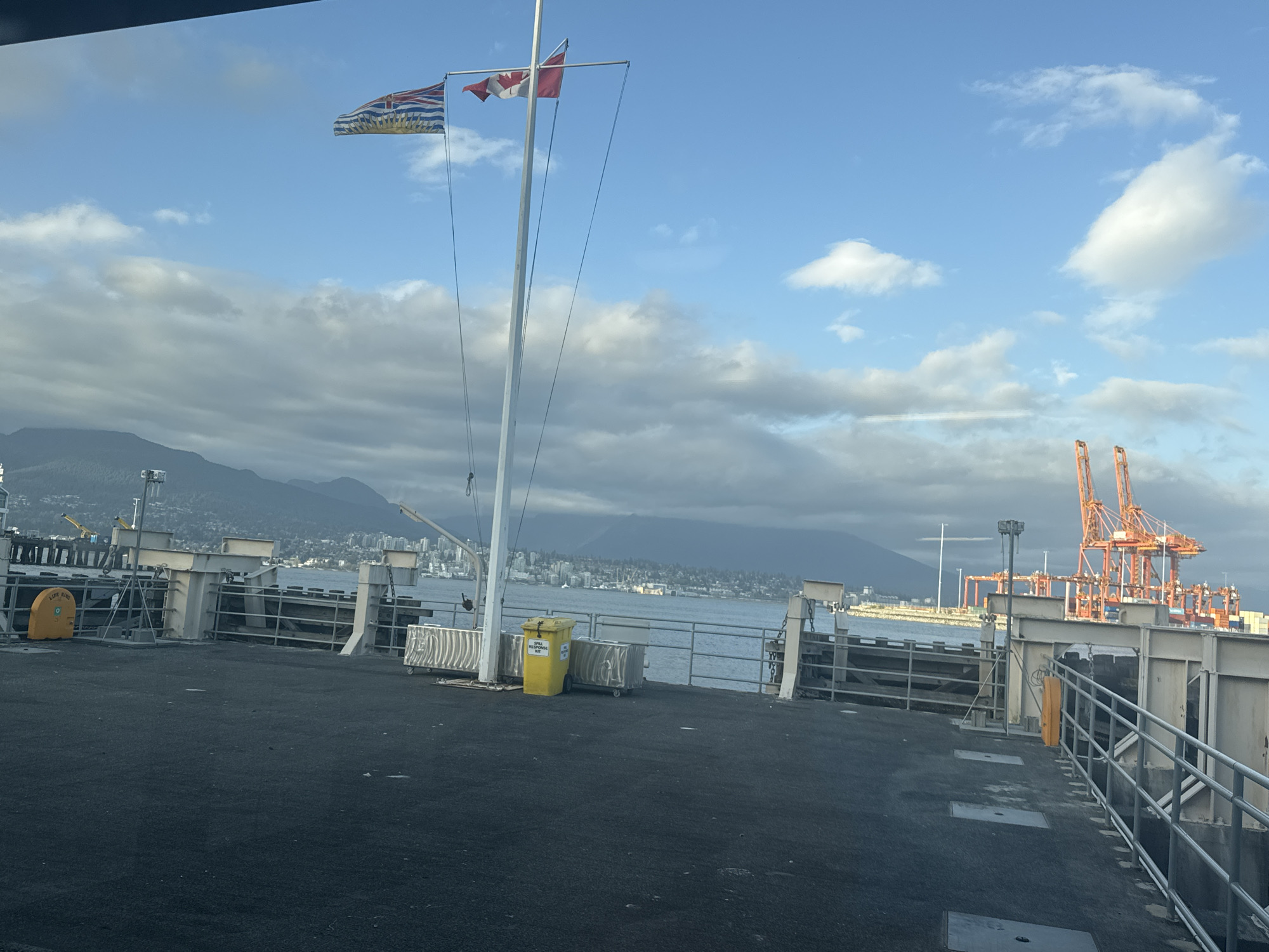 The image shows a view from a pier in Vancouver, Canada, facing towards the city. There is a flagpole with the flag of British Columbia on the left and the flag of Canada on the right. Mountains are visible in the background. In the foreground is a pier surface, with railings and a yellow container labeled "SPILL RESPONSE KIT". In the distance, orange cranes are visible, indicating a port or shipping area. No people are visible in the image. The sky is partly cloudy.