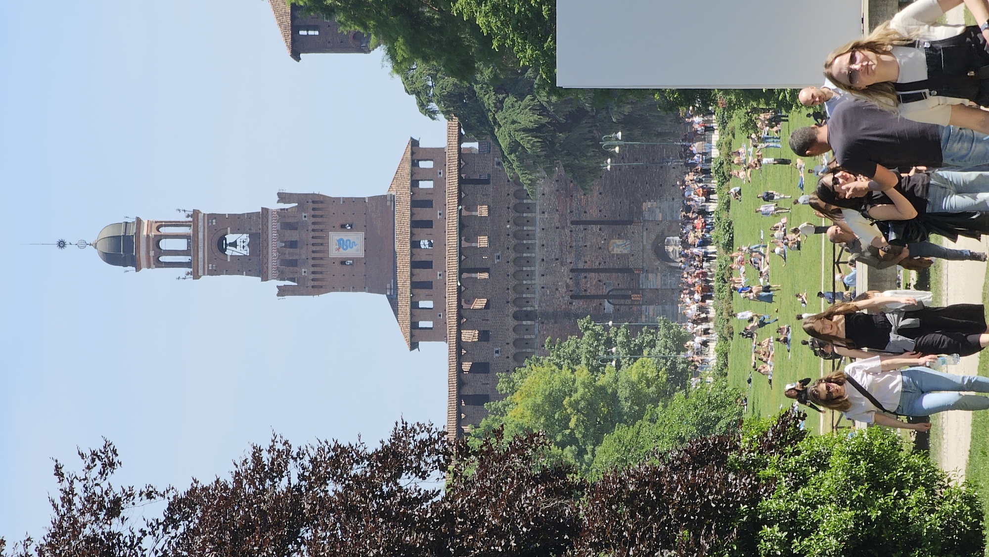 The image captures a sunny day at Sforza Castle in Milan, Italy. The brick structure of the castle dominates the background. A large grassy area is occupied by numerous people, many of whom are lounging and relaxing on the grass. Several individuals stand or walk along a pathway in the foreground. Trees and vegetation partially obscure the lower portion of the castle. The sky is clear blue.
