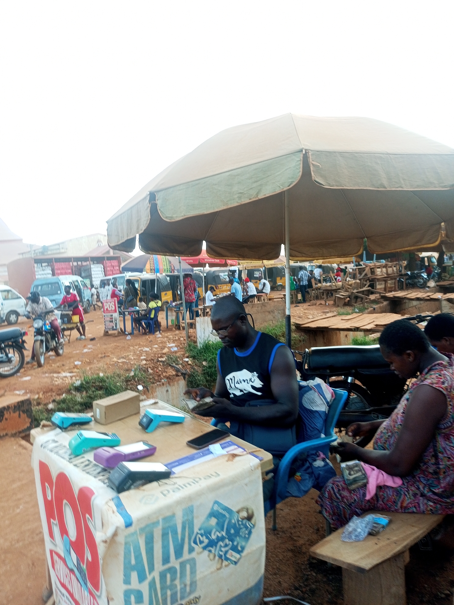In an outdoor setting, possibly a marketplace in Unknown, Unknown, a man wearing glasses and a black tank top with a white logo is seated at a table with multiple point-of-sale (POS) devices. The table is draped with a white cloth printed with "POS ATM CARD." Next to him, a woman in a colorful dress is seated on a wooden bench, counting money. An umbrella provides shade over the area. In the background, there is a bustling scene with people, vehicles, and various stalls. The ground is dirt.