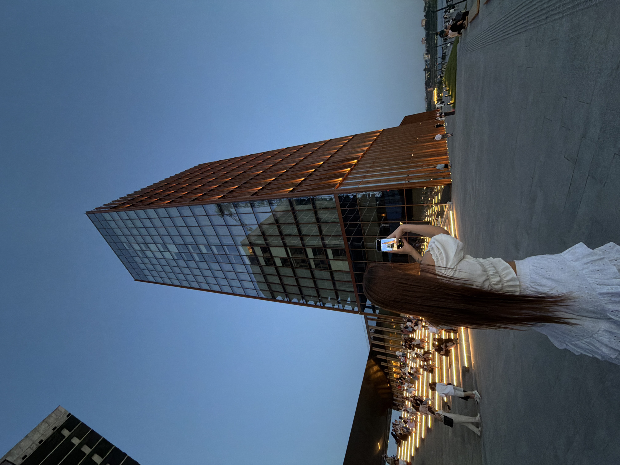 A medium shot depicts a modern glass building with a brown, grid-like exterior, towering against a clear, blue sky at dusk. A young woman with long, brown hair, wearing a white top and skirt, holds up a smartphone, seemingly taking a picture of the building. Below the building, on a concrete surface, are rows of lighted stairs, and people are seated and walking on them. In the distance, along what appears to be a riverbank, there are buildings and vegetation. The location is in the city of Kazan, Russia.