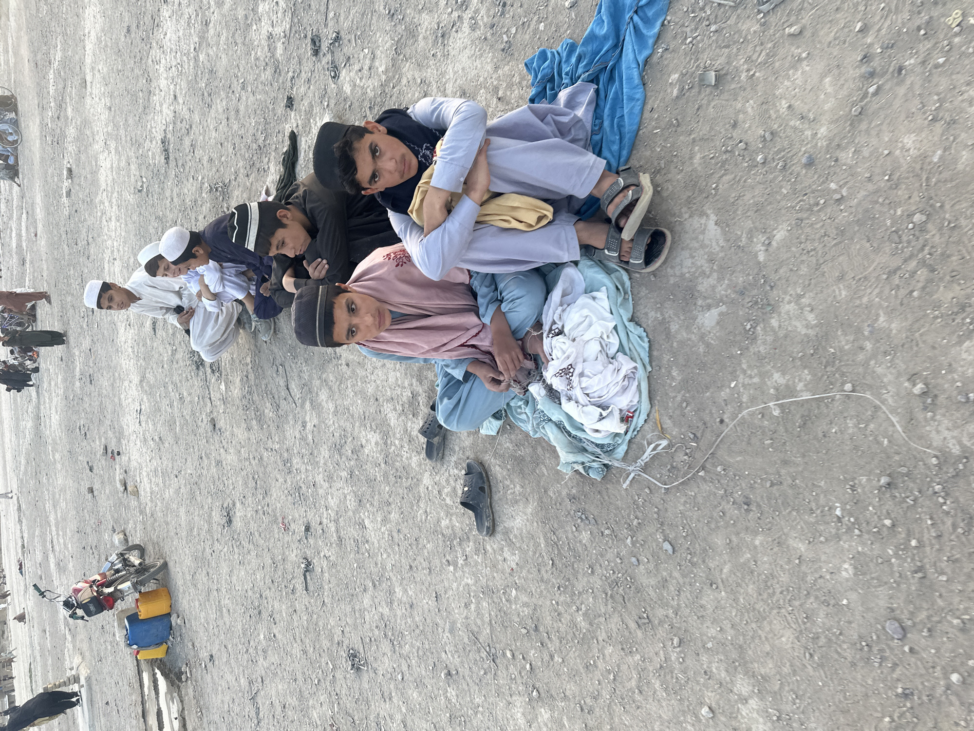 The image appears to depict a group of young males, possibly boys, in Quetta City Tehsil, Pakistan. They are lying on the ground in a row. Some of them wear traditional Muslim clothing and head coverings. Various items, including clothing, sandals, and potentially other belongings are scattered around them on the ground. A motorcycle and supplies are visible in the background. The ground appears to be a rough, outdoor surface.