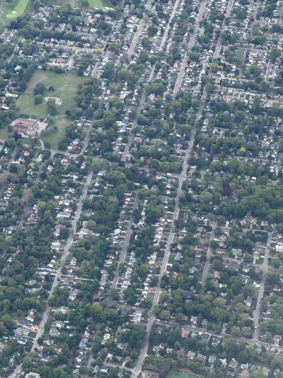 The image shows an aerial view of a residential area, likely in Cresskill, United States, characterized by a grid-like street layout. The scene is dominated by houses of varying sizes and designs, most surrounded by trees and lawns. Vegetation is abundant, giving the area a green and leafy appearance. In the upper left, a golf course is visible. Roads are seen as straight lines intersecting at right angles, typical of planned residential developments. There's also a large brick building next to a park. The overall impression is of a well-established suburban neighborhood.