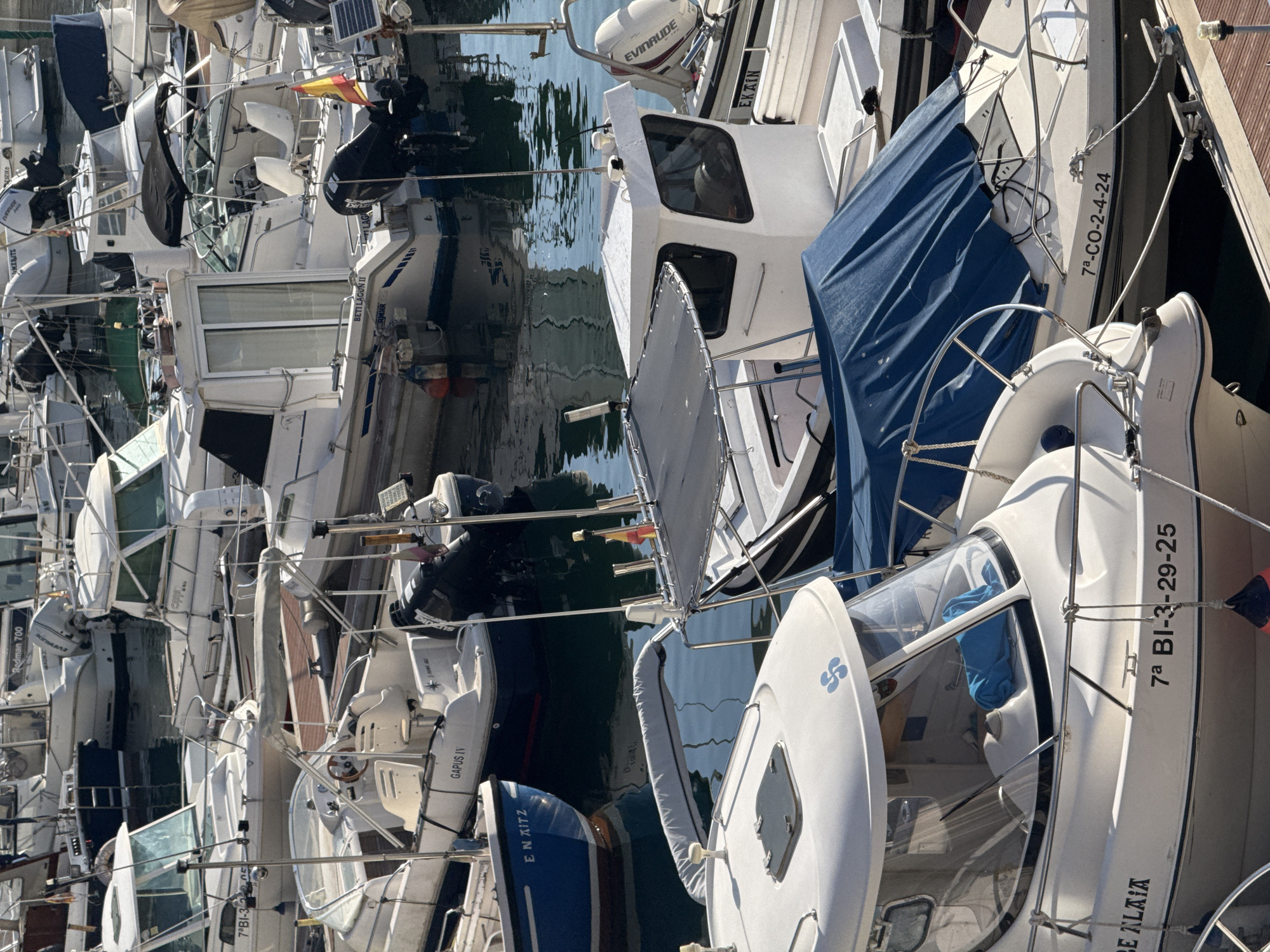 The image shows a harbor filled with numerous boats of various sizes docked together. The boats are primarily white with some dark blue hulls. Several boats have names and registration numbers visible on their sides. One boat in the foreground has the name "BE ALAIA" and the registration number "7ª BI-3-29-25". Another boat has the registration number "7ª-CO-2-4-24". Some boats are covered with blue tarps. The water in the harbor appears calm, reflecting the boats and the sky. The scene is set in Bermeo, Spain, a coastal town known for its fishing industry, as evidenced by the presence of many fishing and leisure boats.