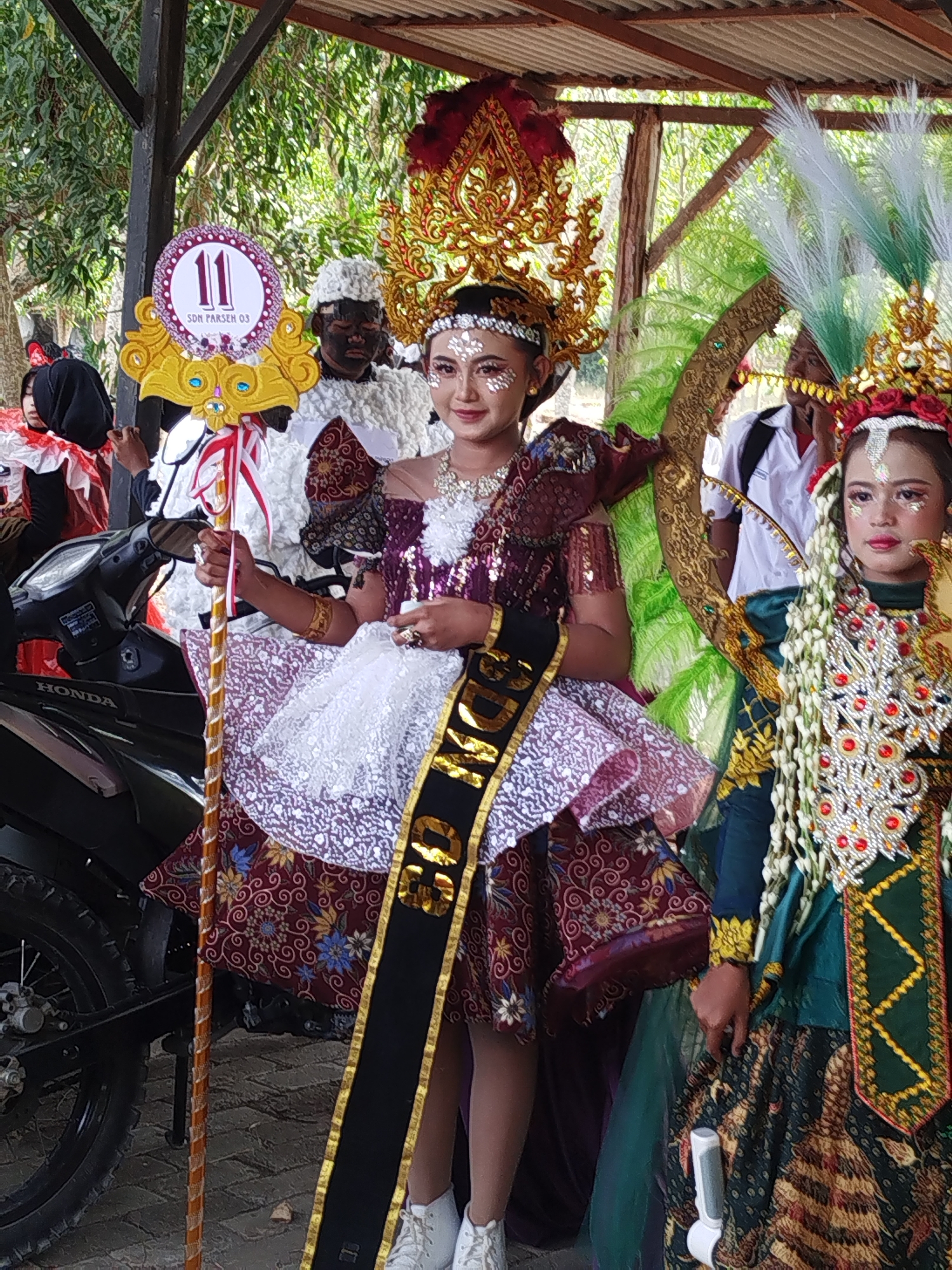 In Jambu, Indonesia, a group of people are participating in what appears to be a parade or cultural event. The central figure is a young woman adorned in a vibrant, ornate costume. She wears a large, golden headdress with red and gold embellishments, and her face is decorated with shimmering body glitter. A black sash with gold lettering, reading "SDN PARSEM 03," is draped diagonally across her chest, and she holds a decorative staff. Her outfit consists of a patterned maroon skirt and a white lace overskirt, with a detailed bodice. To her right, another young woman is also dressed in traditional attire, featuring a green garment adorned with white floral garlands and red jewels, as well as a golden headdress. In the background, a person with their face painted black and wearing a white, fluffy costume is visible. A black Honda motorcycle is parked to the left of the central figure. The setting appears to be outdoors, possibly under a covered structure with a corrugated roof, with trees and foliage visible in the background.