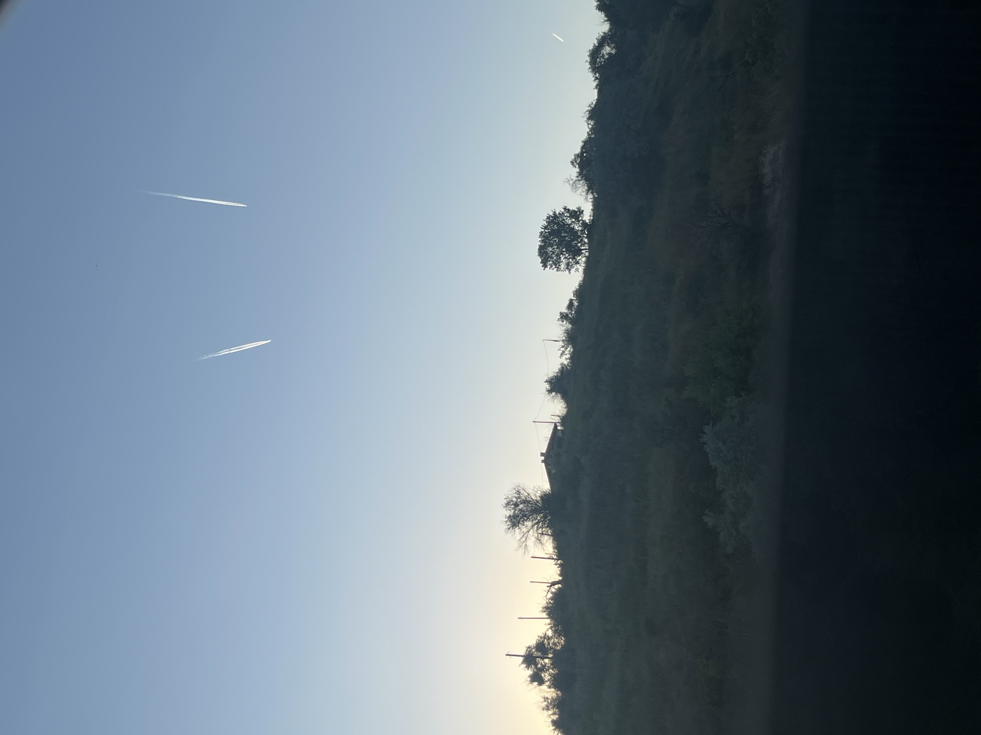 An outdoor, horizontal, medium shot captures a rural landscape in Varna, Bulgaria, during twilight. The sky, a gradient of pale blue to a lighter hue near the horizon, features two distinct white contrails from aircraft. Below the sky, a silhouetted hilly terrain is covered in vegetation and scattered trees. A small building with a reddish roof is visible atop the hill, partially obscured by trees and vegetation. The foreground is dominated by a dark, out-of-focus area, possibly a road or vehicle. No people or animals are visible in the frame.