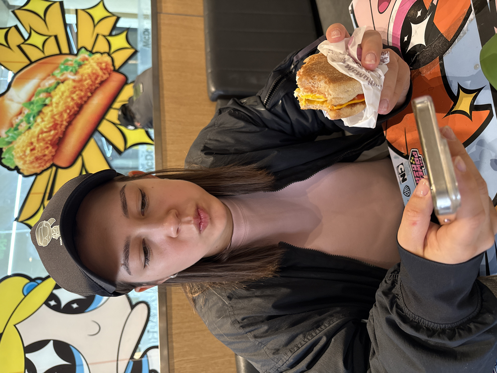 A woman is sitting in a McDonald's restaurant, holding a phone in her right hand and a breakfast sandwich in her left. She is wearing a black baseball cap, black jacket, and a light pink top. The table has a Powerpuff Girls design. A McDonald's advertisement featuring a sandwich is visible in the background. This photo was taken in the city of Dongcheng District, China.