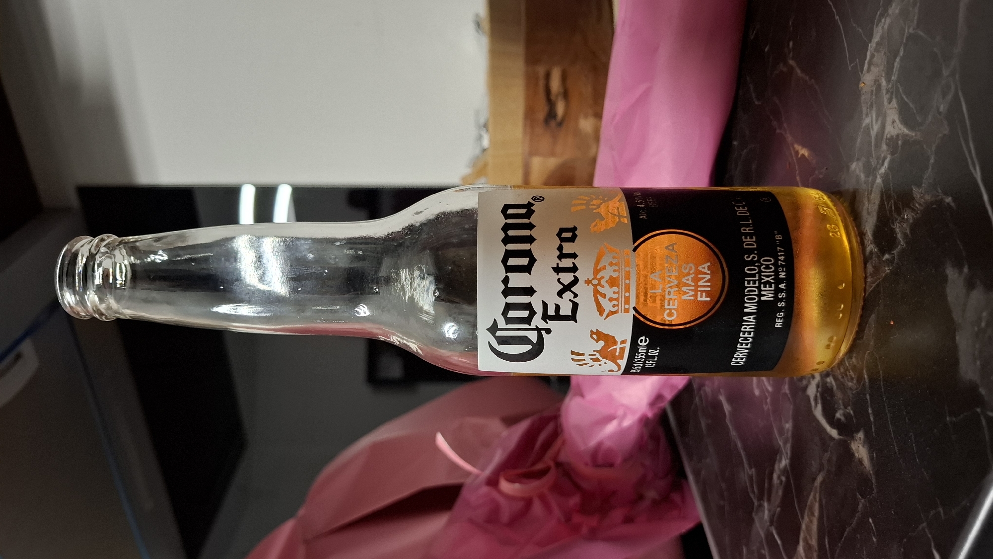 A clear glass bottle of Corona Extra beer is shown, centered in the frame, standing upright on a dark, marble-patterned surface. The bottle is mostly empty, with a small amount of golden-colored liquid at the bottom. The label on the bottle is black and orange, featuring the "Corona Extra" logo in white stylized text, along with a crown and a circular emblem containing text. To the left of the bottle, a crumpled piece of bright pink fabric is visible, partially obscuring the background. The background consists of a dark, out-of-focus area and a lighter, softly lit vertical surface. The scene appears to be indoors. The location is not Plovdiv, Bulgaria.