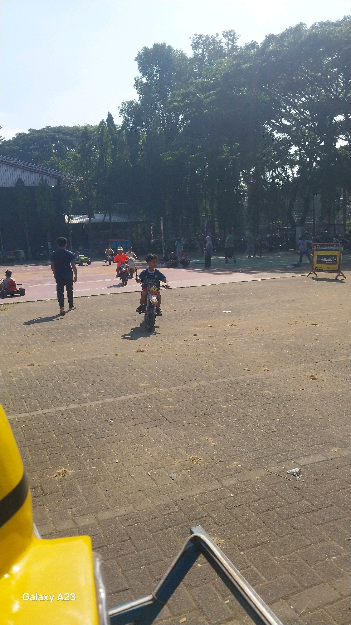 A bright, sunny outdoor scene unfolds on a paved area in Malang, Indonesia, likely a park or recreational space. The foreground is dominated by a yellow object with a black stripe, possibly part of a ride or attraction, with a blue metal frame visible to its right. Beyond this, a young boy is the central focus, riding a small dirt bike with a yellow and black design. He is wearing a dark t-shirt with white writing. To his left and slightly behind him, a man in a dark t-shirt and pants stands with his back to the camera, casting a long shadow.
In the middle ground, other children are also on small motorbikes. One child wears an orange shirt and another a yellow shirt. Further back, a group of people are gathered, some sitting on the ground, others standing. A large building with a metal roof is visible behind them, with a wooden-like structure to its left. The background is filled with lush, tall trees, suggesting a well-vegetated area. A yellow sign with black writing, possibly advertising, is propped up on a stand on the right side of the scene. The ground is paved with rectangular bricks, with scattered dry leaves and debris. The sky is clear and bright blue.