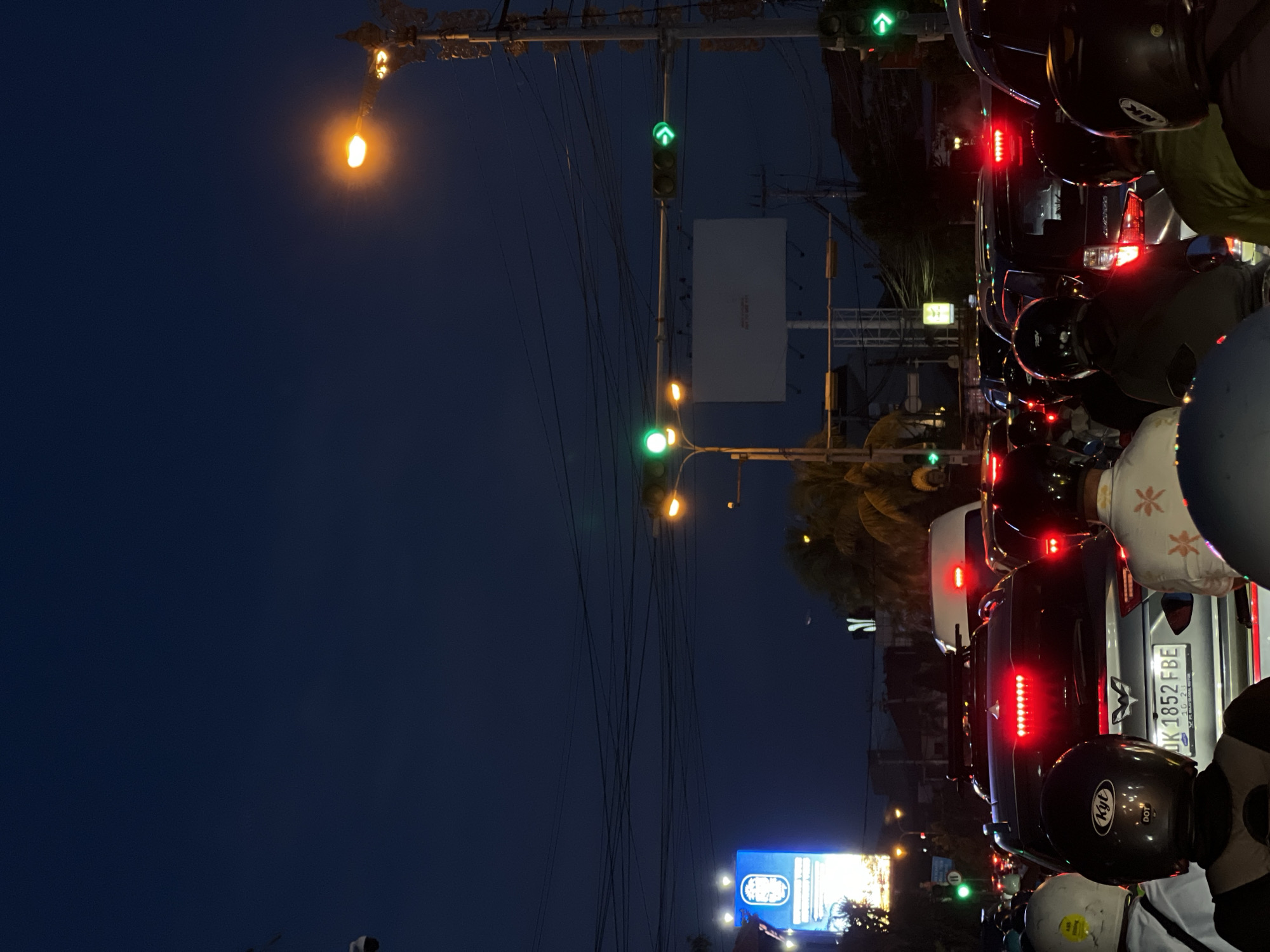 The image, originally rotated 90 degrees clockwise, depicts a dense urban street scene at night in Denpasar, Indonesia. The dark blue sky signifies the late evening or nighttime setting, illuminated by various artificial light sources.
In the foreground and extending into the mid-ground, a queue of vehicles, predominantly motorcycles and cars, is halted in traffic. Numerous bright red brake lights are illuminated, indicating the vehicles are stopped. Several individuals, likely drivers or passengers on motorcycles, are visible from behind, wearing helmets.
Overhead, a dense entanglement of utility wires stretches across the frame. Multiple traffic lights are visible; notably, one displays a green upward arrow, signaling permission to proceed for certain traffic directions, while others are simply green. Streetlights emit a warm orange glow, contributing to the ambient lighting.
In the background, a large, unadorned white billboard stands, and further away, a vibrant blue illuminated sign with Asian script is visible. Tropical foliage, possibly palm trees, can be discerned, hinting at the geographical location. A visible car license plate reads "DK 1852 FBE," confirming the location as Bali, Indonesia (DK is the regional code for Bali), with "10-21" also present, likely indicating an expiration date. A "Kyt" brand helmet with "DOT" certification is also clearly seen. The scene captures the typical atmosphere of a busy Indonesian city street after dark.