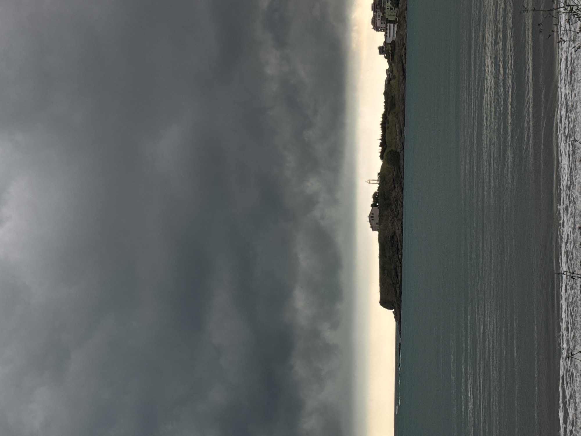 The image captures a dramatic coastal scene under a heavily overcast sky, likely in Tsarevo, Bulgaria. The upper portion of the frame is dominated by expansive, dark grey clouds, suggesting either an approaching storm, a very cloudy day, or the low light of dawn or dusk. A narrow, brighter band of light is visible along the horizon, creating a striking contrast with the dark clouds above and indicating the primary source of illumination.
Below the sky, a rugged coastline or headland stretches horizontally across the image. This elevated land is sparsely covered with dark vegetation. Several buildings, including some white structures and others with visible roofs, are perched on the higher ground overlooking the sea, possibly residential or vacation properties. A distinct, small white structure, possibly a monument or part of a navigational beacon, is visible on a lower section of the headland.
The lower part of the image features the sea, which appears in a deep blue-green color, reflecting the muted light. The water's surface is textured with numerous small ripples, indicating subtle movement. On the far right, a narrow strip of foamy waves breaks onto a dark, possibly sandy or pebbly beach. Two fishing rods are clearly planted upright in the wet sand near the water's edge, suggesting recreational fishing, though no people are visible. The overall mood is solemn and atmospheric, with the dark clouds hinting at dramatic weather conditions. There is no visible text in the image.