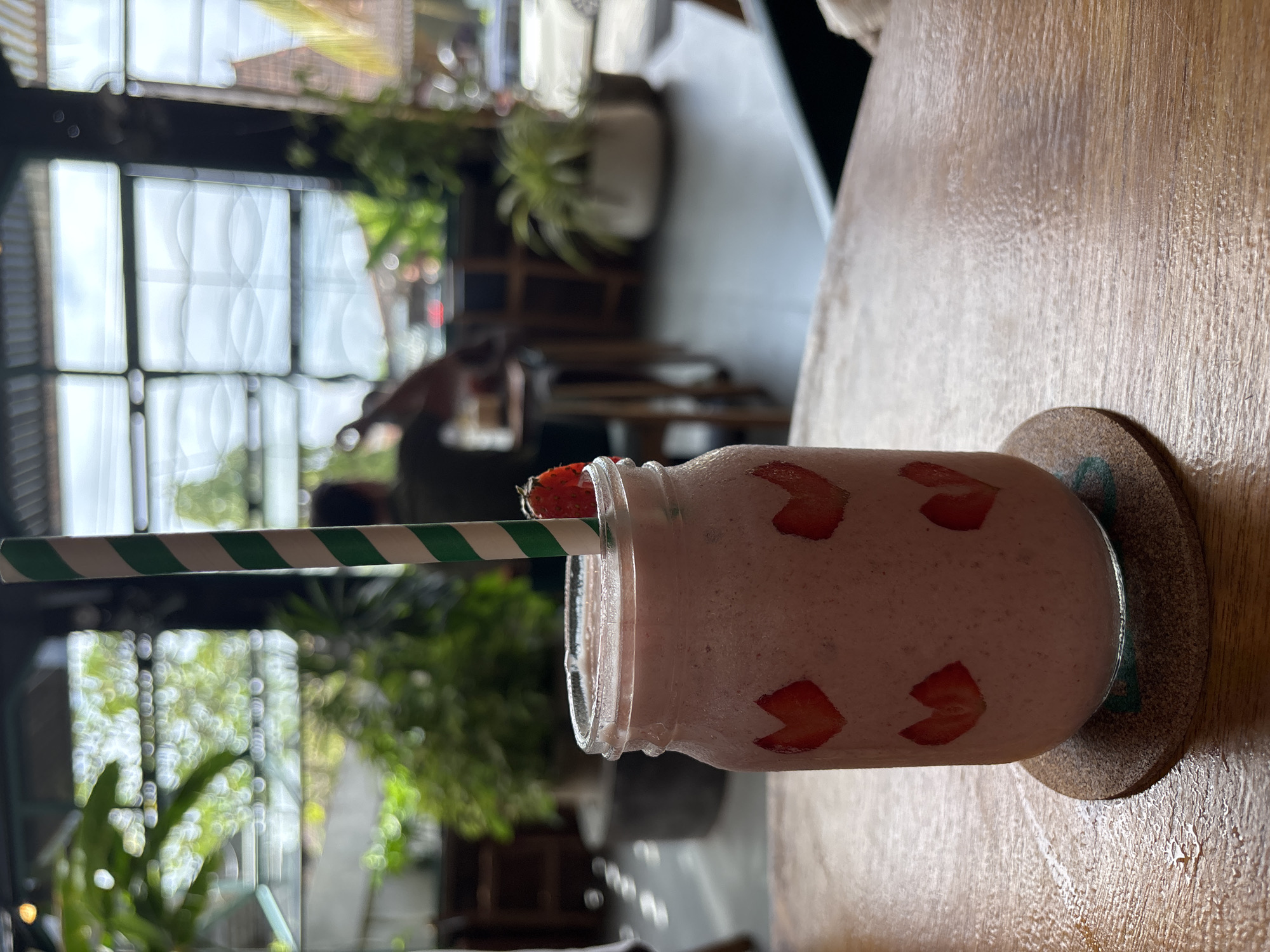 A tilted eye-level shot depicts a strawberry smoothie in a glass jar with sliced strawberries affixed to the exterior. A green and white striped paper straw rests through a strawberry slice on the jar's rim. The jar sits on a round, brown coaster on a wooden table. The background reveals an indoor area with large windows, greenery, and diffused natural light. Through the windows, a building is seen across the street. The setting suggests an outdoor seating area of a cafe, likely in Mengwi, Indonesia.