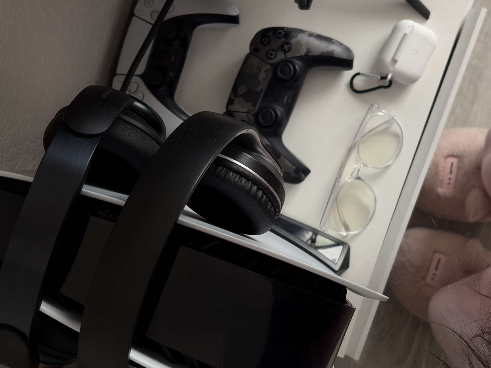 The image shows a close-up of a PlayStation 5 console with black headphones resting on top. Next to it are two PlayStation controllers (one white, one camouflage), a pair of clear-framed eyeglasses, and a white AirPods case with a black carabiner. To the right, a pair of pink slippers is partially visible. The setting appears to be an indoor surface, possibly a desk or shelf. The location in Saint Petersburg, Russia is not discernible from the image.