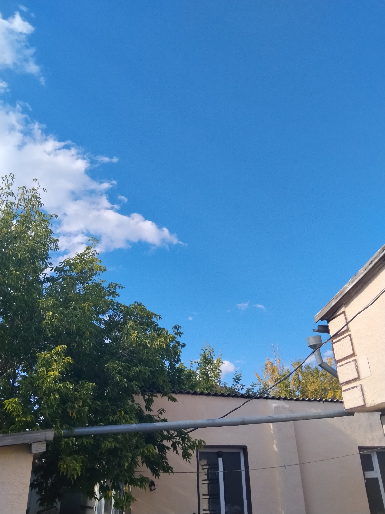 The image is a low-angle shot of a building's exterior with a bright blue sky and scattered white clouds above. Green trees with dense foliage are visible on the left side of the frame, partially obscuring the building. A long, grey pipe runs horizontally across the middle of the image, supported by the building. The building itself is light-colored with visible windows and architectural details along the upper edge. A downspout is attached to the side of the building. The scene is likely in an outdoor urban environment, possibly a residential or commercial area in Kokshetau, Kazakhstan, given the context. There are no people or discernible actions depicted in the image.