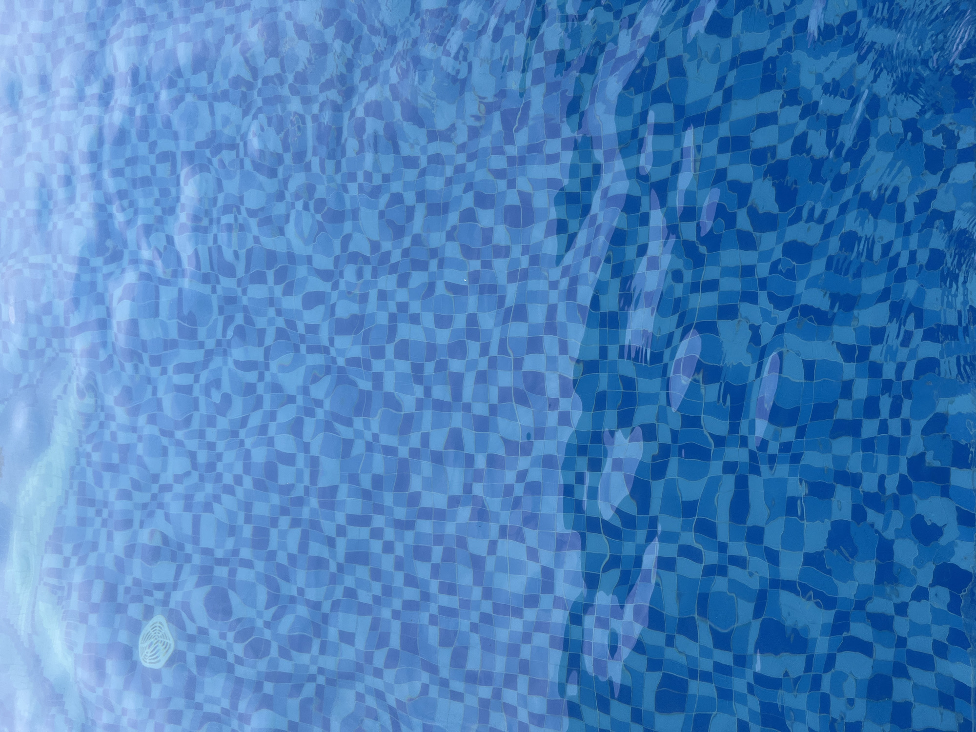 The image shows the tiled bottom of a swimming pool. The tiles are a mosaic of varying shades of blue, creating a checkered pattern. The surface of the water is disturbed by ripples, distorting the appearance of the tiles beneath. In the upper left quadrant of the image, a white circular pool skimmer is visible just beneath the water's surface. The scene is of a swimming pool, likely in a resort or residential area, but there is no specific visual information in the image to pinpoint its location within Bac Nha Trang Ward, Vietnam. No people or actions are depicted.