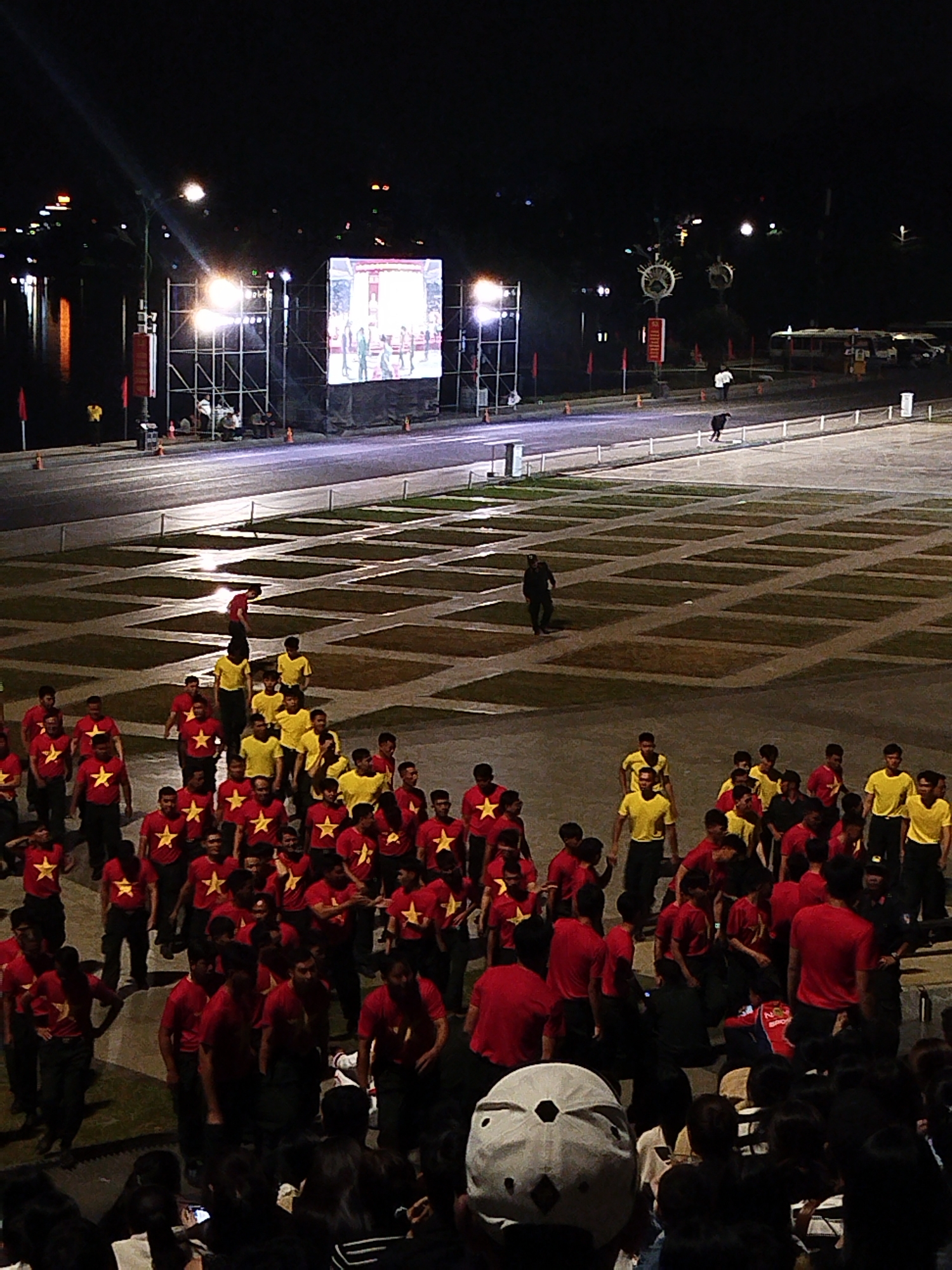 This media file captures a nighttime outdoor event in Da Lat, Vietnam. A large crowd of people, mostly young males, are gathered in a plaza. Many are wearing red shirts with a yellow star, and some are wearing yellow shirts. The setting includes a large outdoor screen displaying images, a stage with scaffolding and lighting equipment, and a body of water in the background. The ground is paved with a grid pattern of dark squares.
The activity appears to be some kind of assembly or performance, with groups of people marching or standing together. Several individuals are walking across the plaza, including some in dark clothing, possibly security personnel or organizers. The atmosphere seems to be one of anticipation or engagement with the event.
Notable details include the bright artificial lighting from the stage and screen illuminating the scene, contrasting with the dark night sky. The presence of an audience in the foreground suggests this is a public event. The attire of the participants, particularly the red shirts with yellow stars, is a strong visual cue that could indicate a nationalistic or team-oriented event. The time of day is clearly night. No specific weather conditions are discernible.
There is some text visible on the screen, but it is not legible enough to read. A red flag with yellow markings is also visible on a pole near the stage.